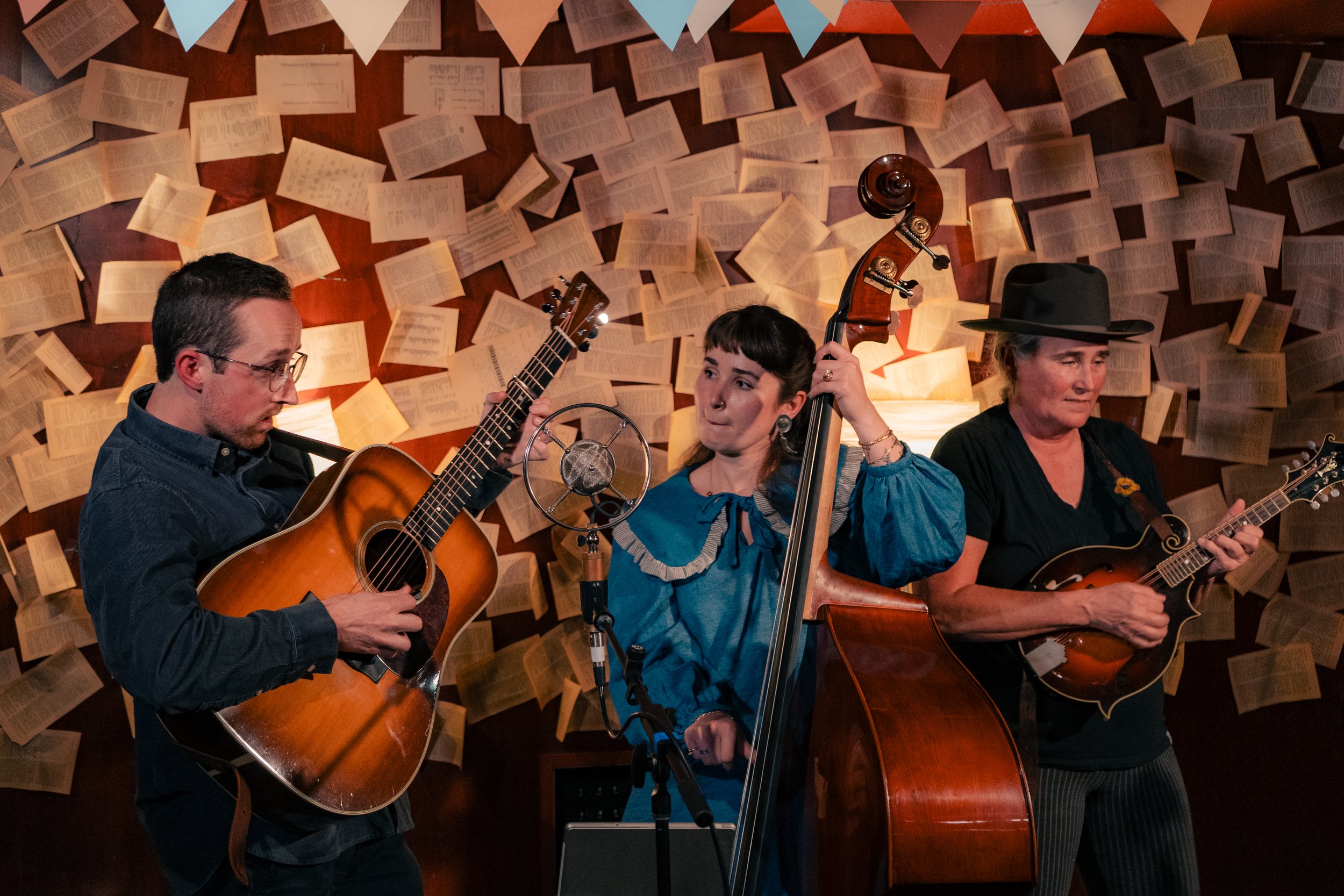 Three musicians performing with guitars and a double bass in front of a wall decorated with open books and hanging strings of colorful pennant banners.