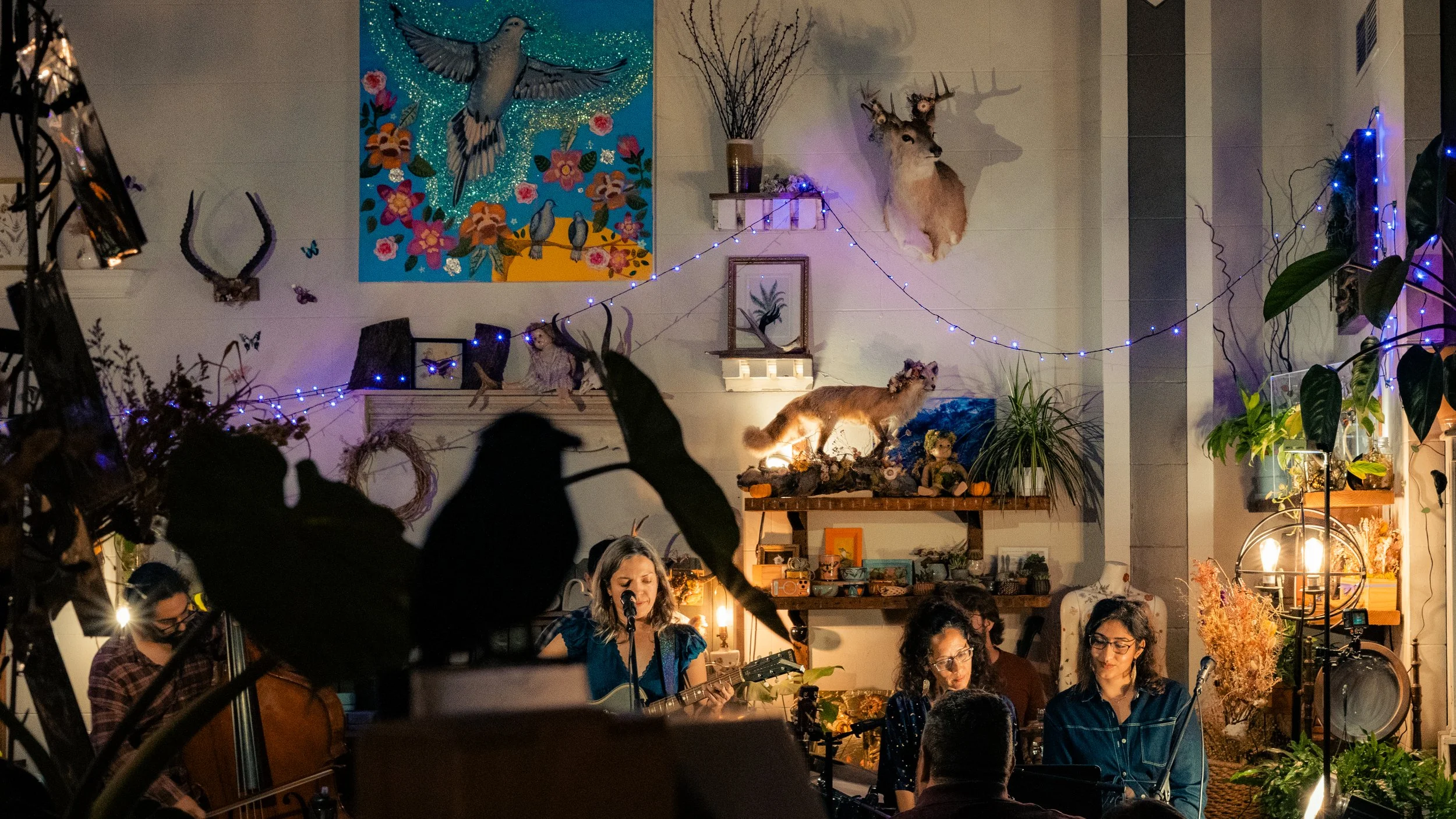 People performing music in a cozy, decorated indoor space with plants, string lights, and animal-themed decorations, including mounted deer and antelope heads, and paintings on the walls.