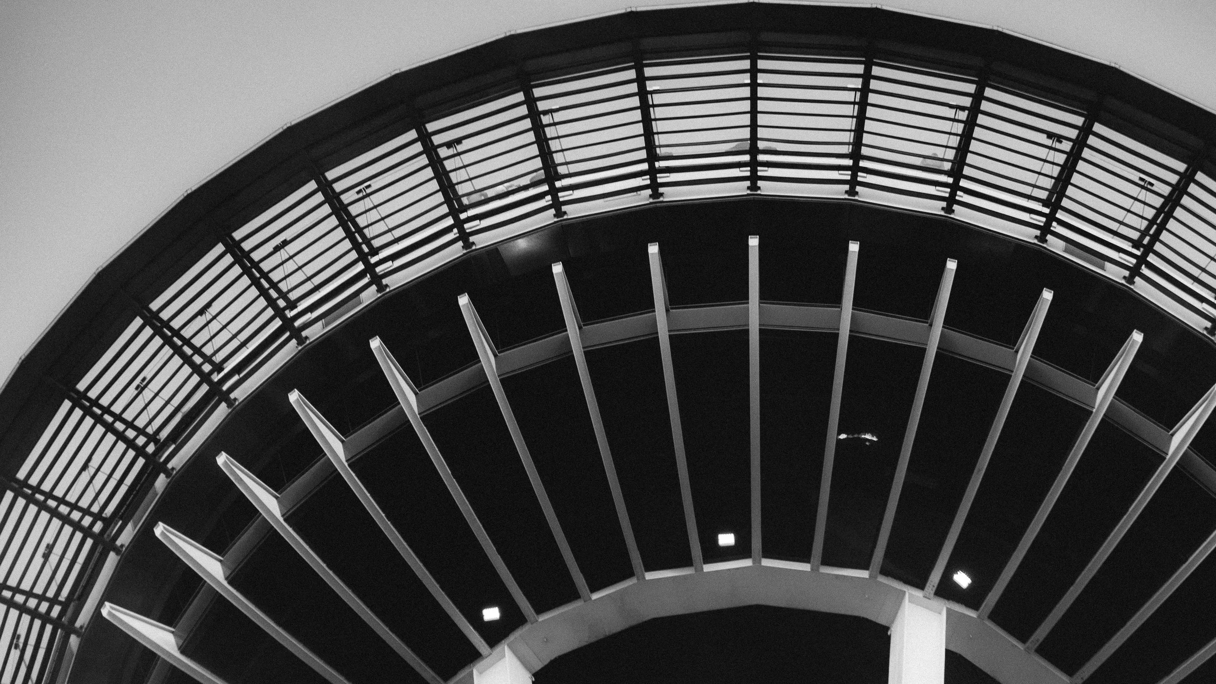 A modern building with an arched design, featuring horizontal and vertical metal slats and a curved upper balcony with railing, captured in black and white.