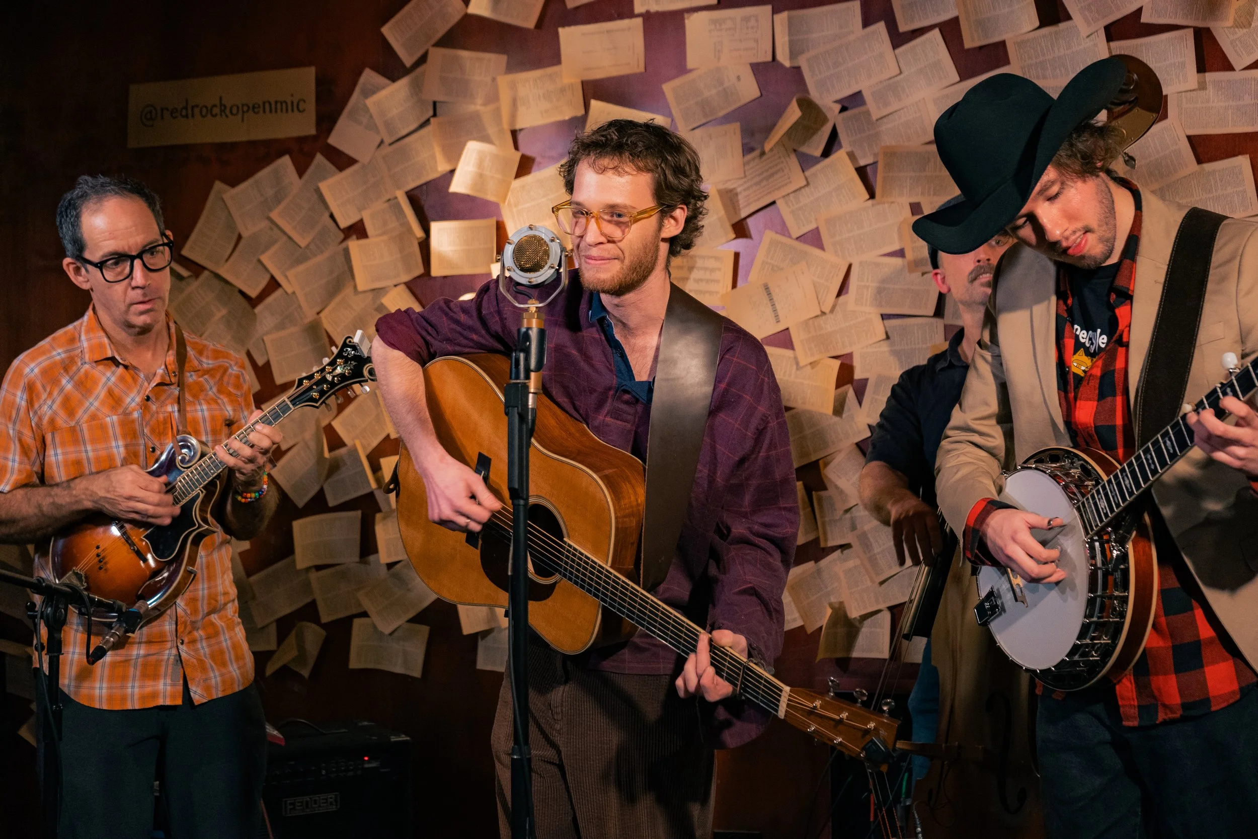 A group of four musicians performing in a room decorated with pages from books on the wall; two are playing guitars, one is playing a banjo, and one is singing into a microphone.