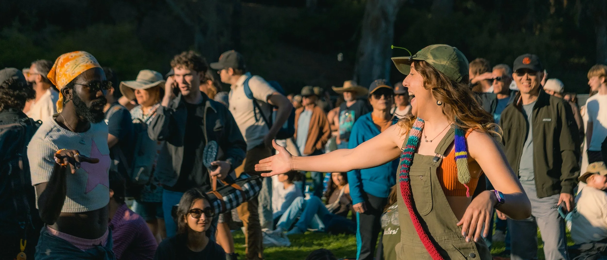 A woman in a green hat and overalls smiling and reaching out to shake hands with a man wearing a bandana and sunglasses at an outdoor gathering with many people.