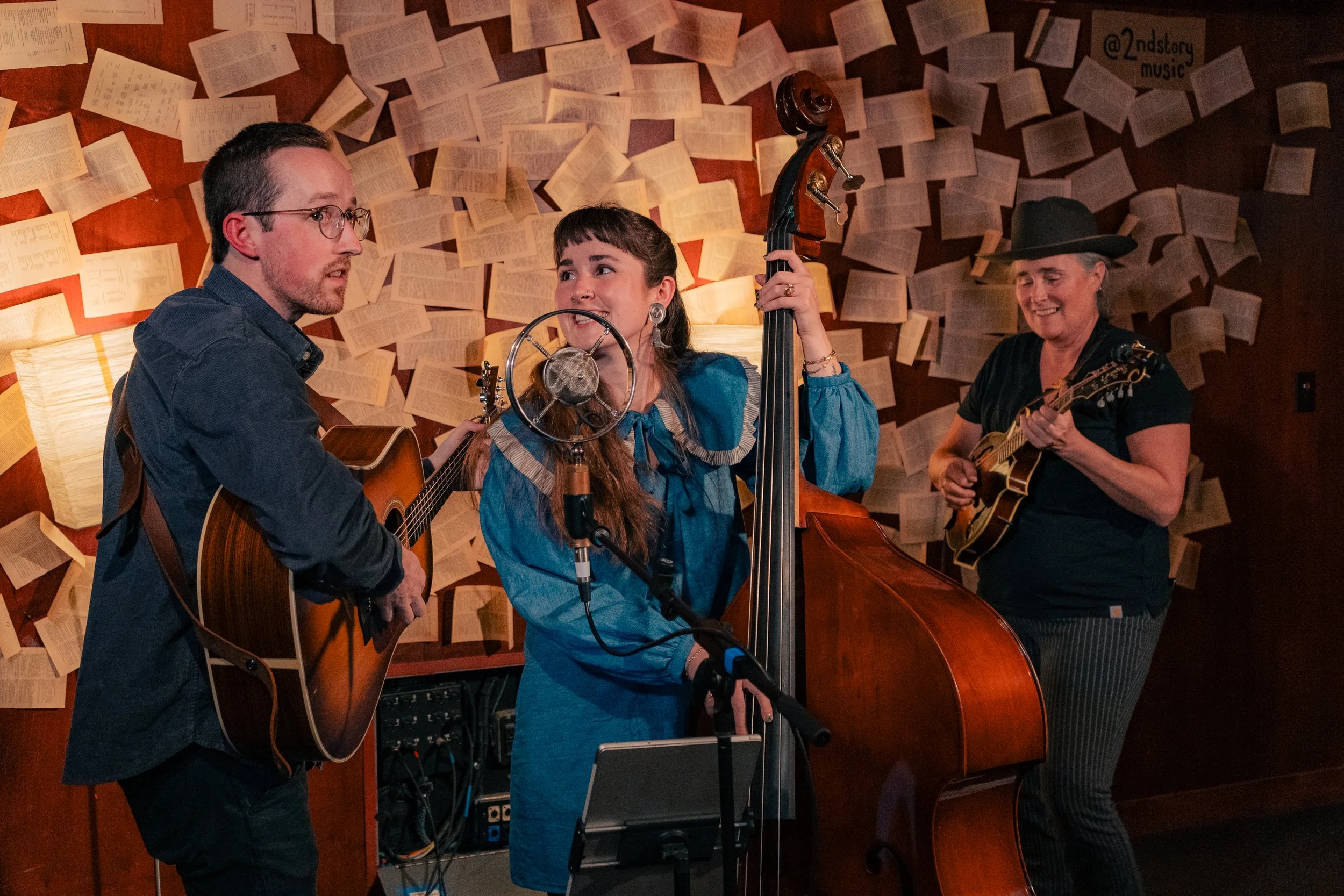 Three musicians performing in a cozy venue: a man with glasses playing an acoustic guitar, a woman with long hair singing into a vintage microphone and playing an upright bass, and another woman wearing a black hat strumming a mandolin. The backgroun