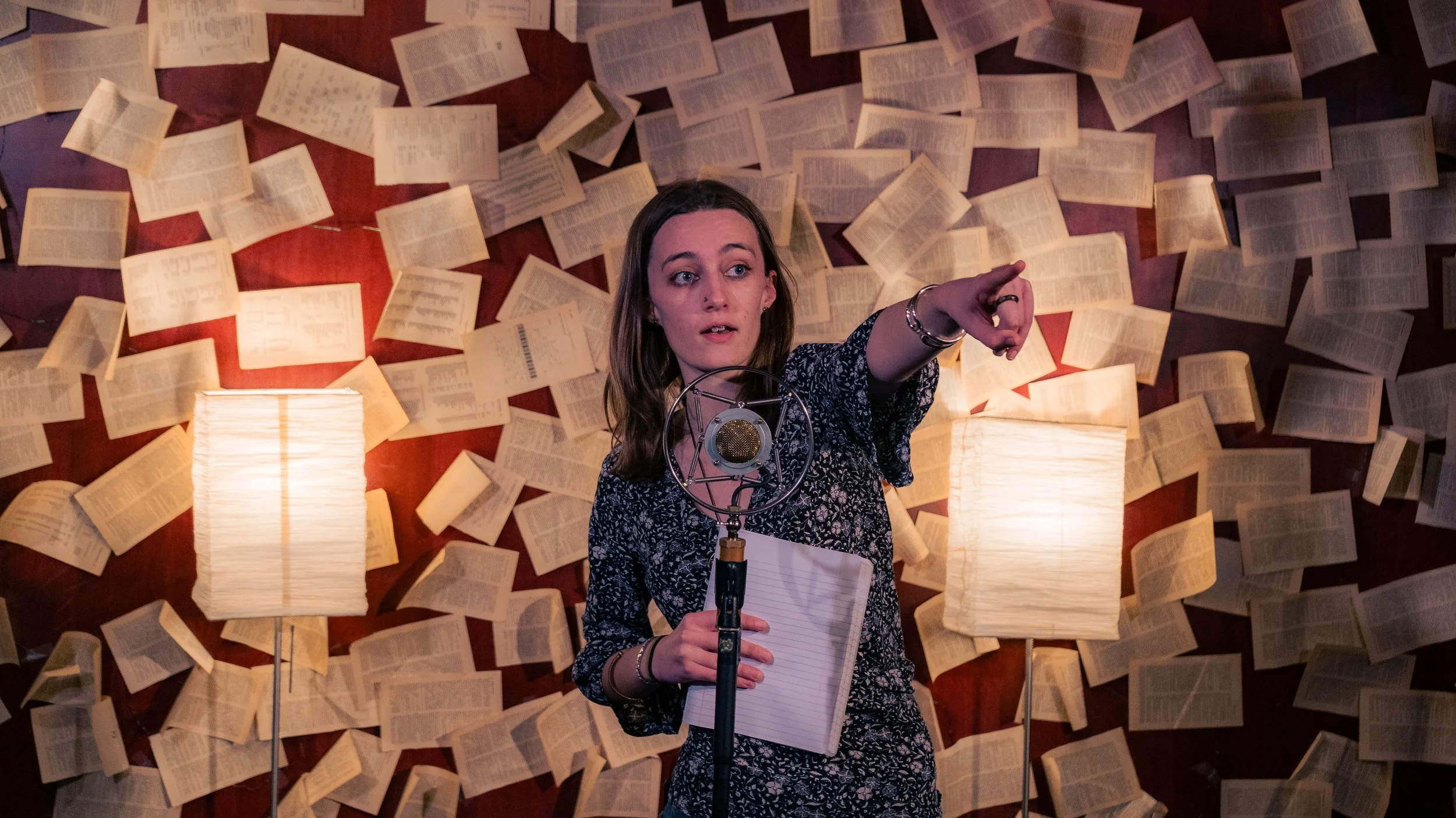 A woman with long brown hair, wearing a dark patterned shirt, stands in front of a microphone with an open notepad in her left hand. She is pointing to her right with her right hand. The background features a collage of open books on a wall and two l
