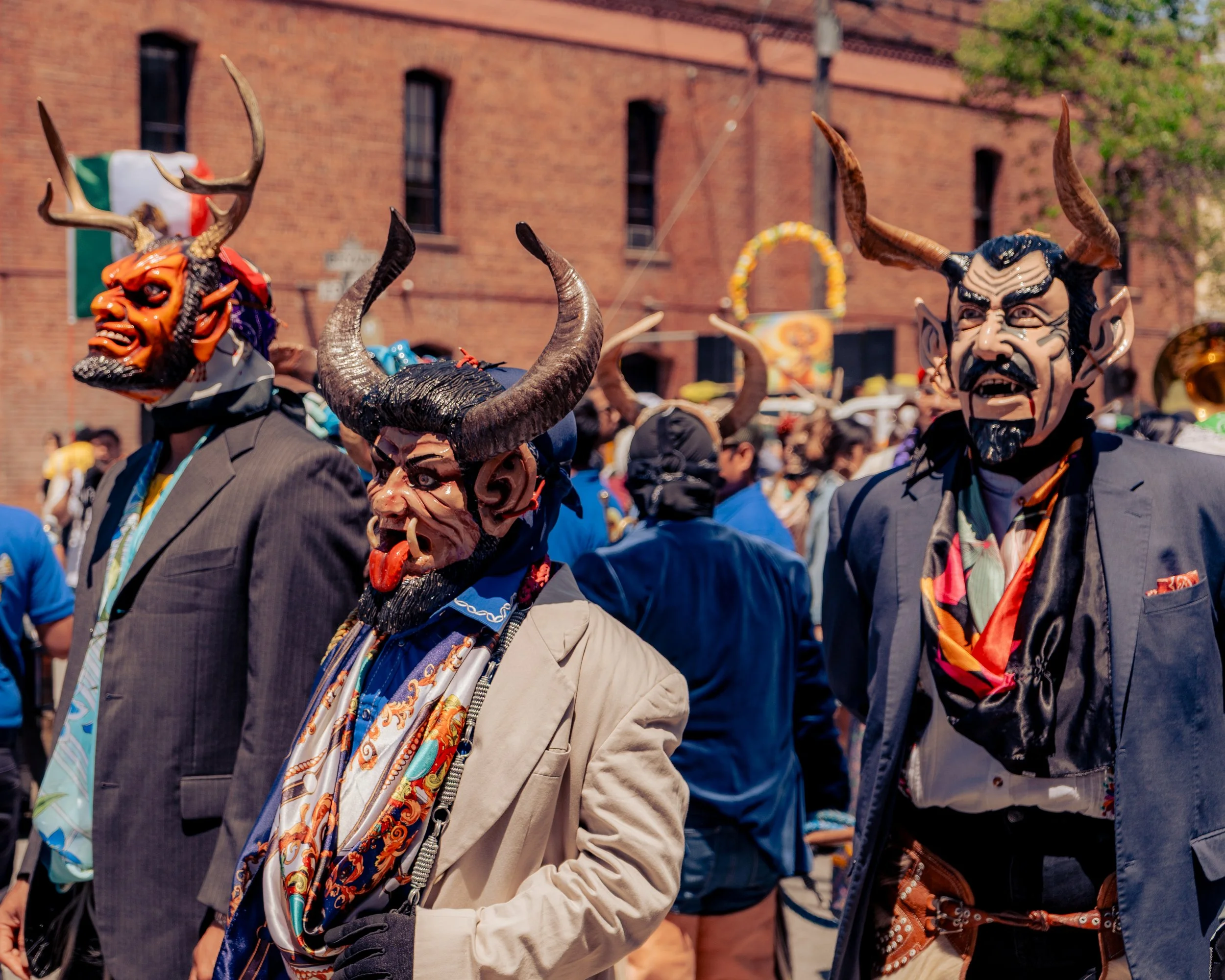 People wearing colorful masks with horns and demonic features, dressed in suits and costumes, participating in a street carnival or parade with a brick building and colorful decorations in the background.
