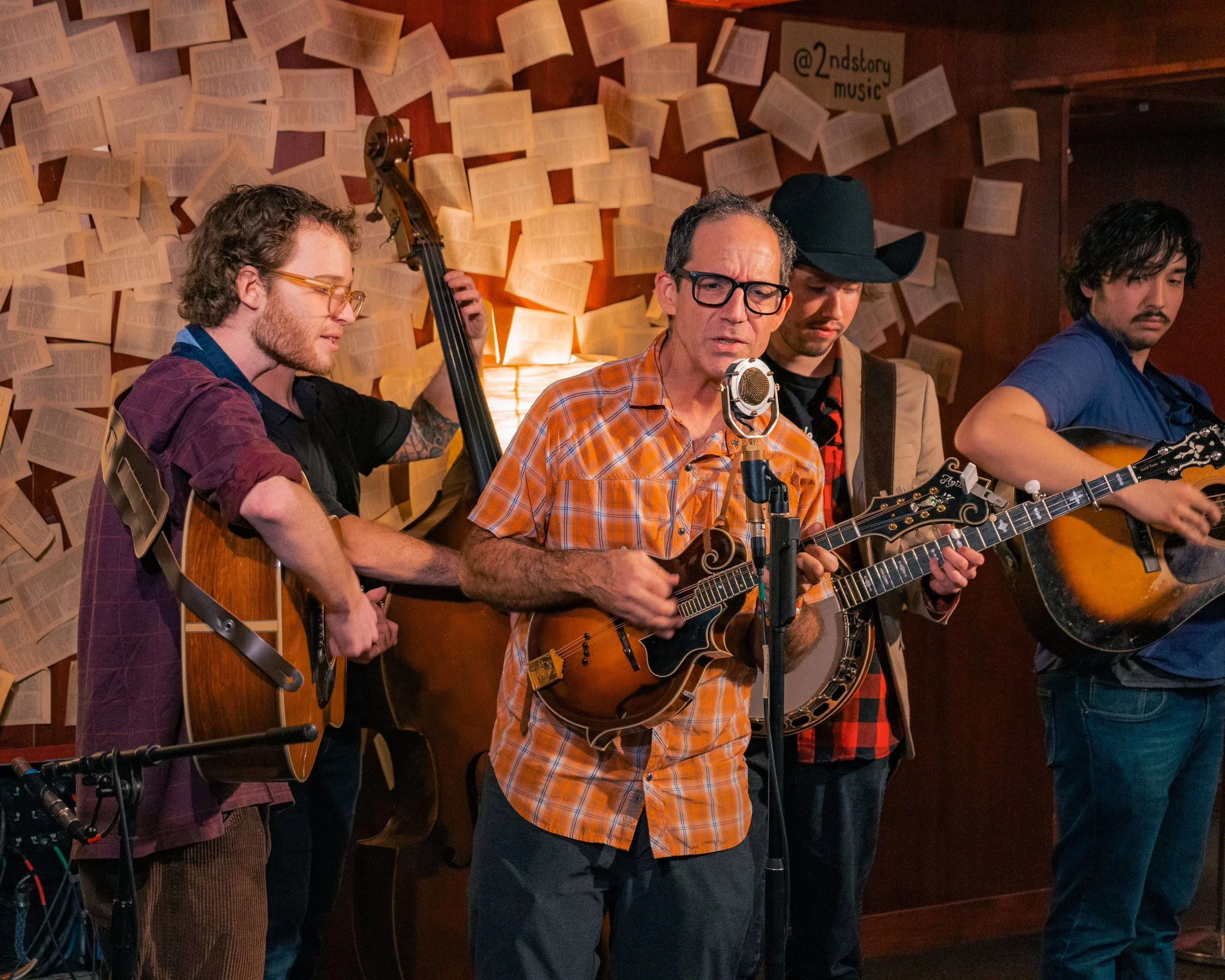 A group of four musicians playing guitars and a banjo during a live performance in a cozy venue with walls decorated with open books.