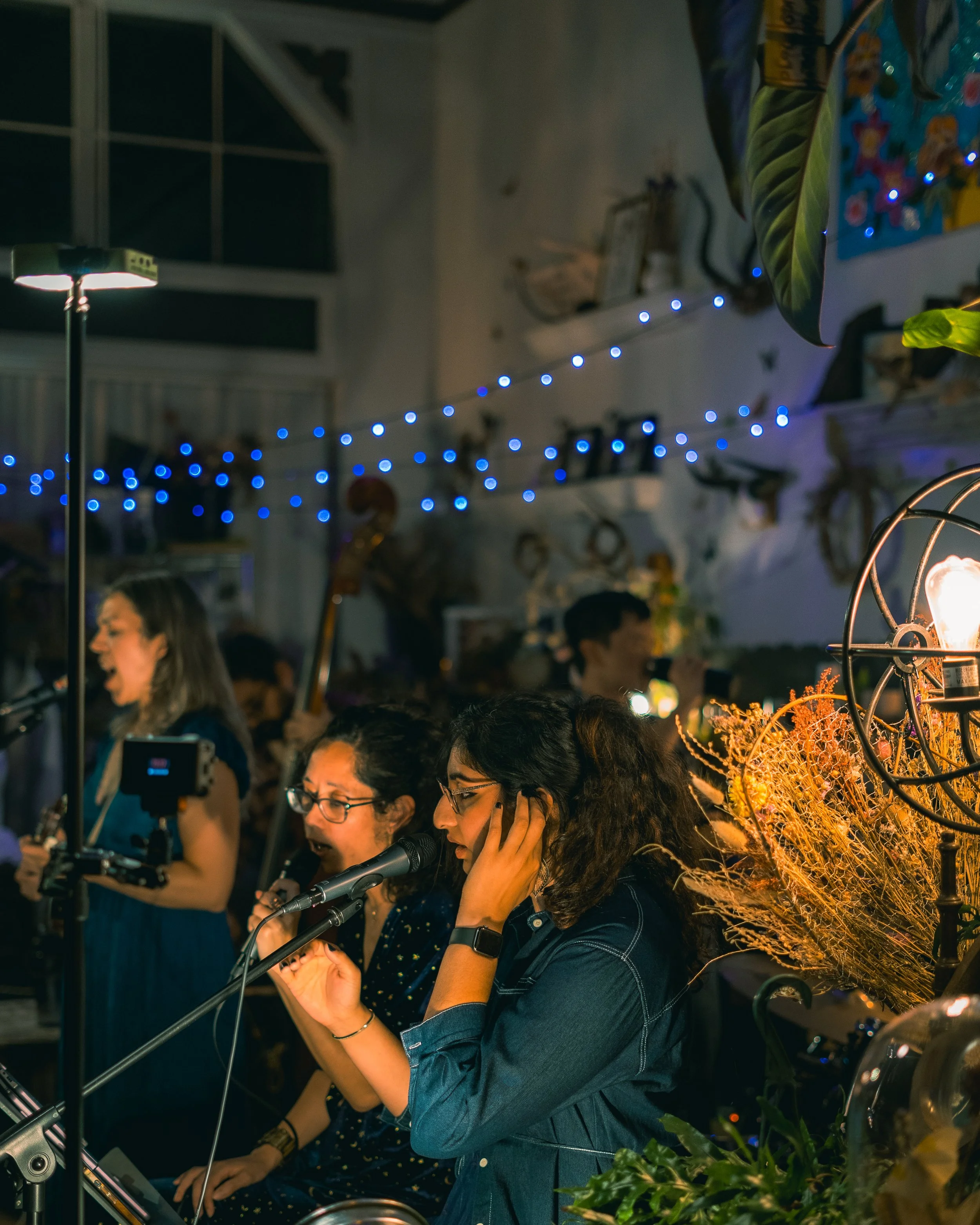 People singing and playing instruments at a live music event decorated with blue string lights and warm lighting.