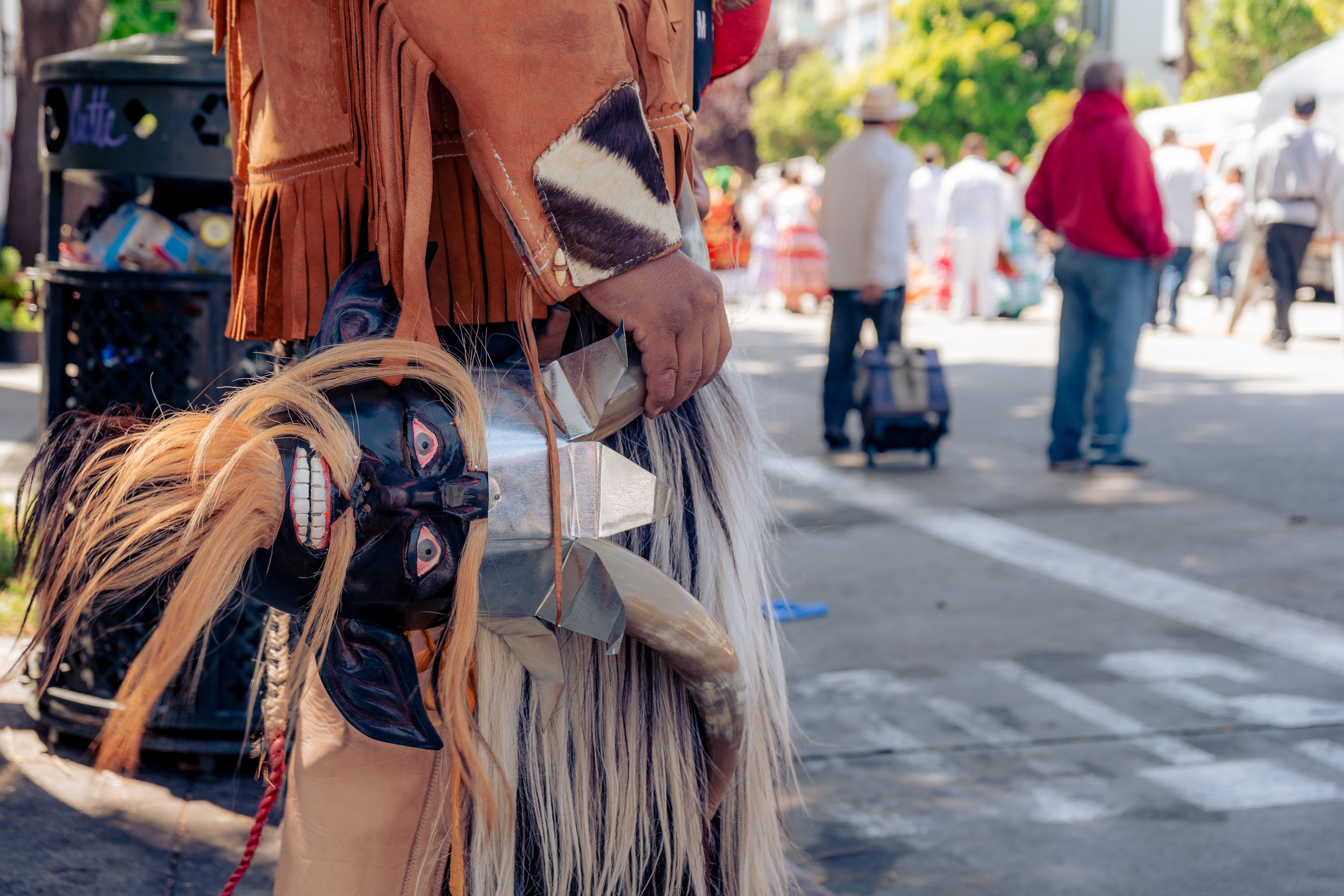 A person holding traditional Native American masks and a staff at a marketplace. People walking in the background with trees and market stalls.