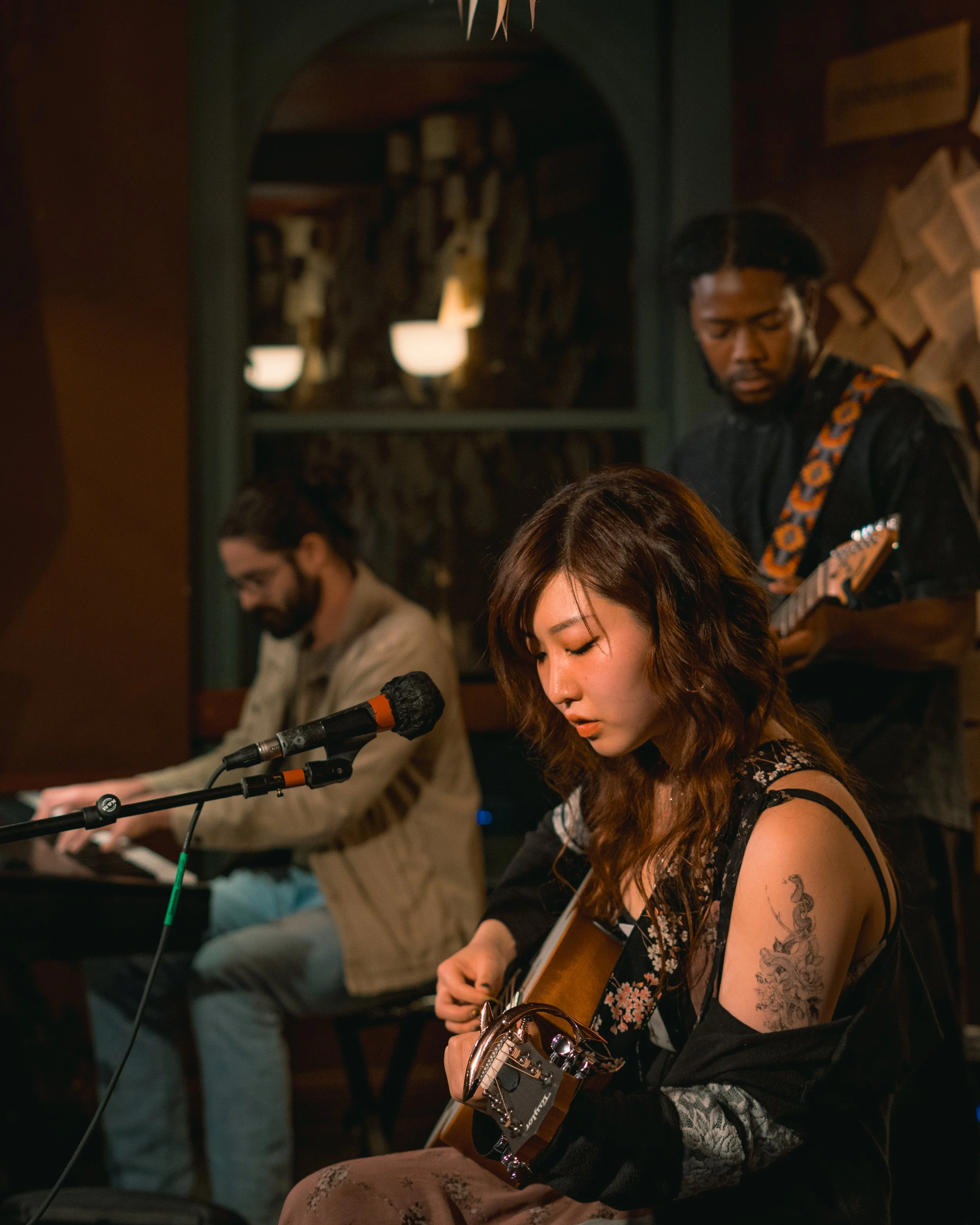 A woman with shoulder-length wavy hair playing an acoustic guitar, with a tattoo on her upper arm, in a dimly lit cozy music venue. Two men, one on a keyboard and another playing an electric guitar, are in the background, blurred.