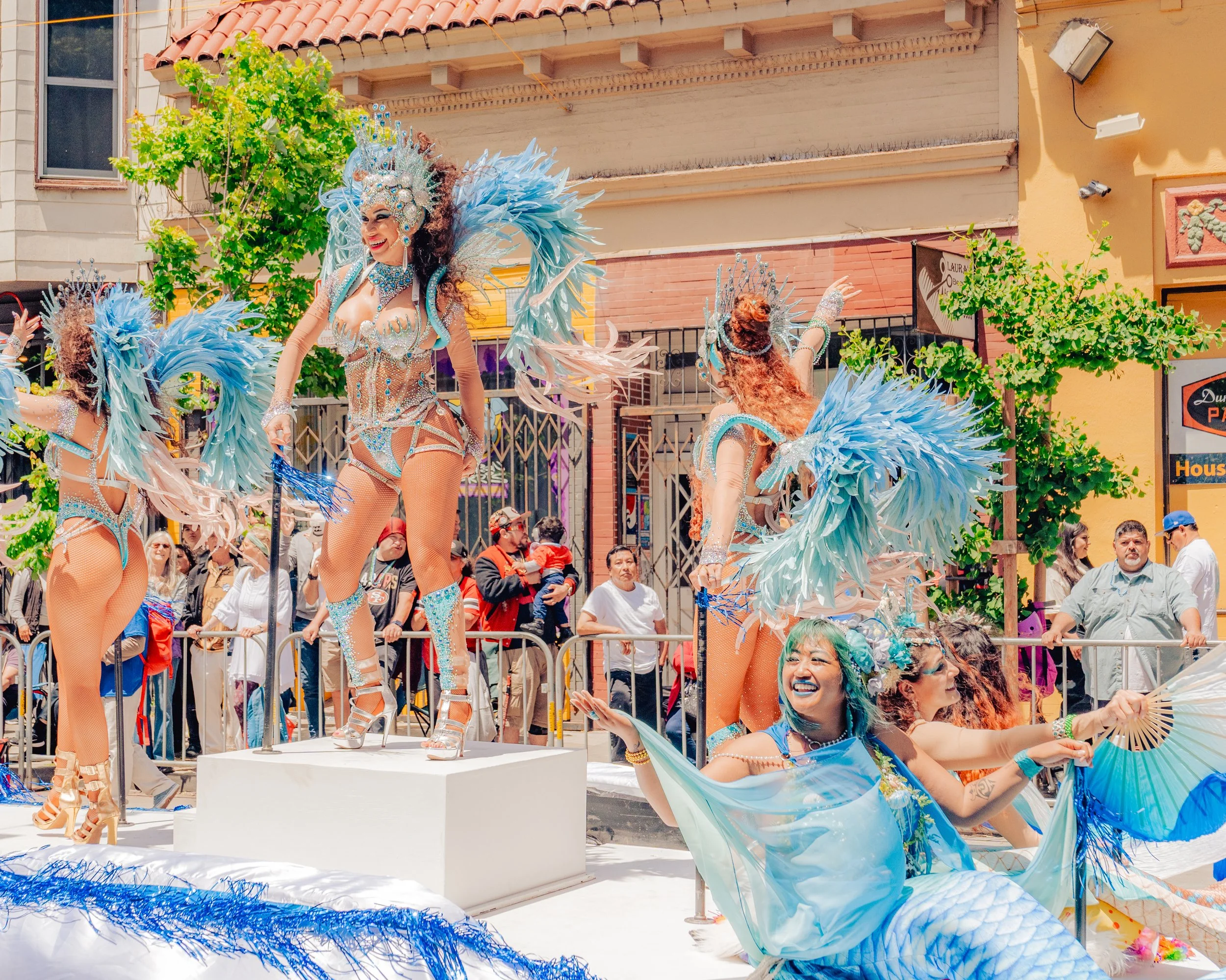 Parade float featuring performers in elaborate blue and silver costumes with feathers and sequins, dancing and posing on a decorated platform during a daytime parade.
