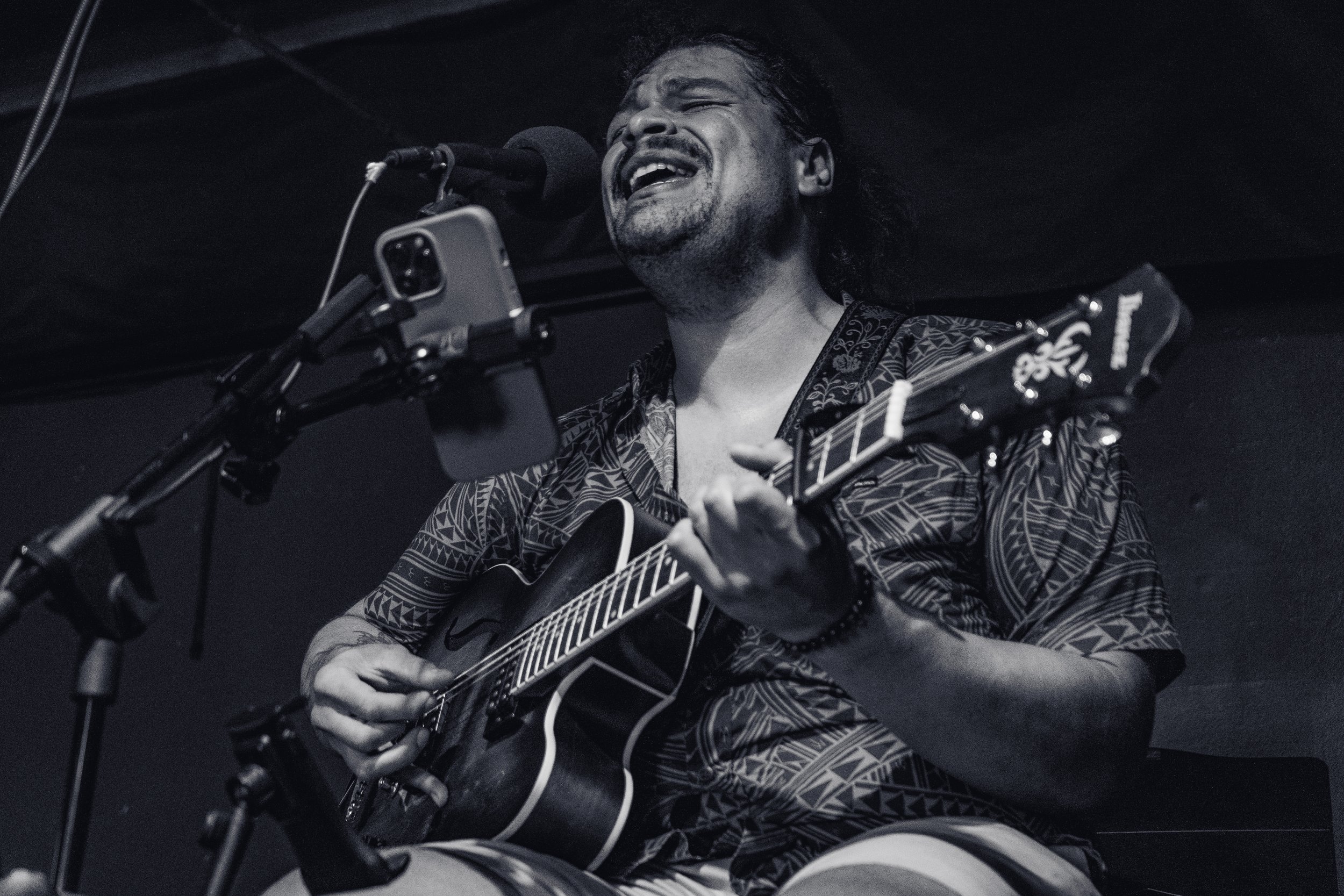A man with long hair, tattoos, and a patterned shirt is passionately singing into a microphone while playing an acoustic guitar in a dark setting.