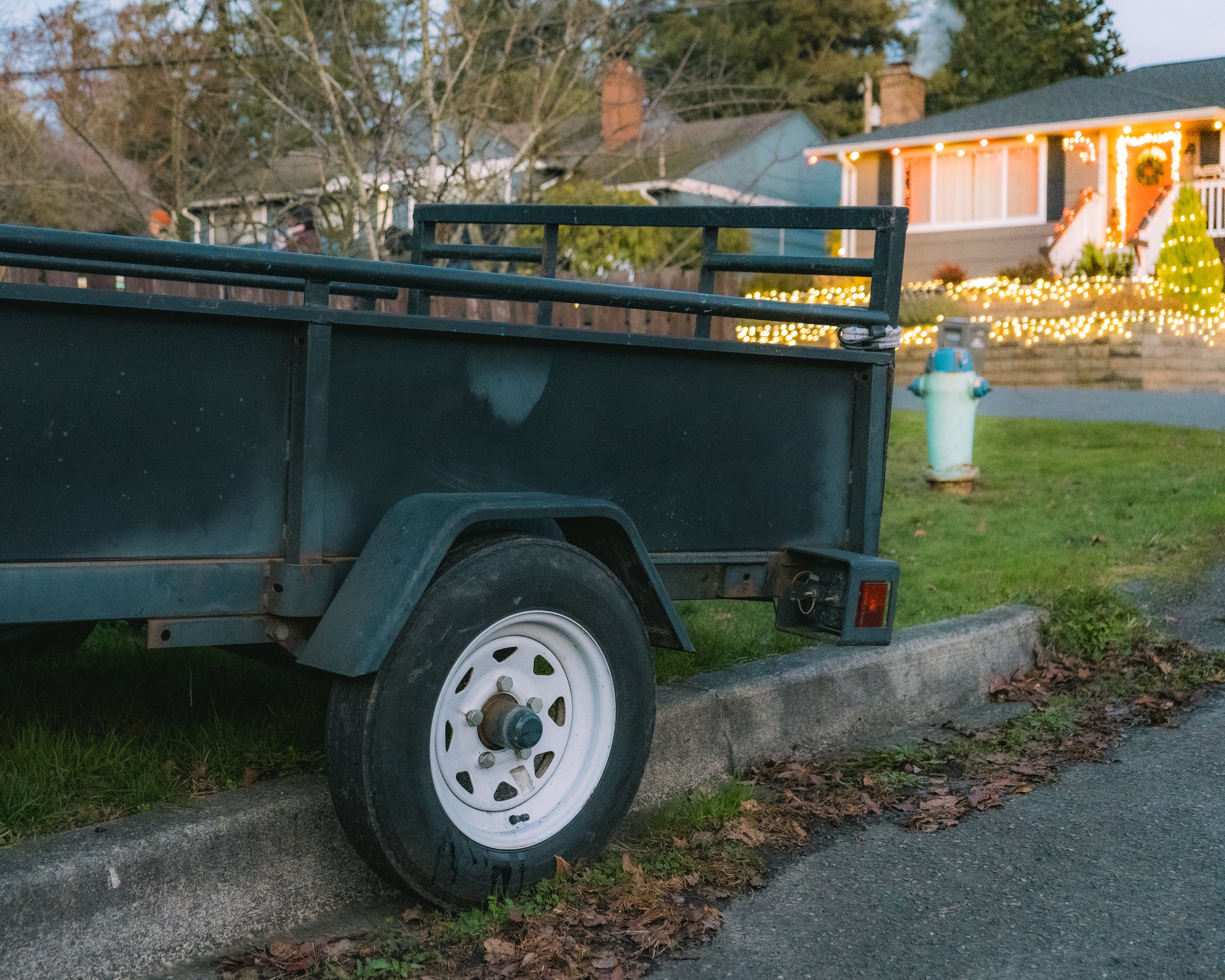A black utility trailer parked on a curb with a house decorated with Christmas lights in the background.