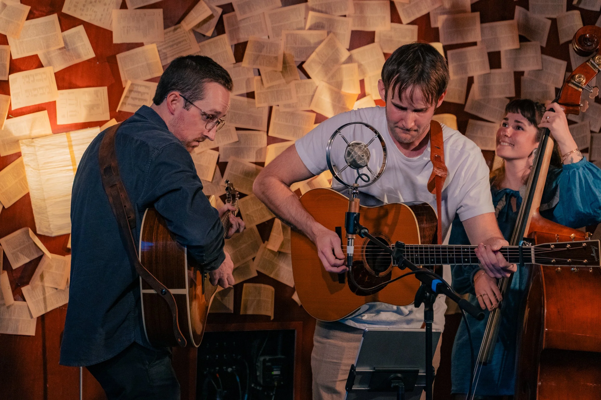 Three musicians playing guitars and a double bass in a cozy, intimate setting with a wall decorated with open books and a lamp, creating a warm ambiance.