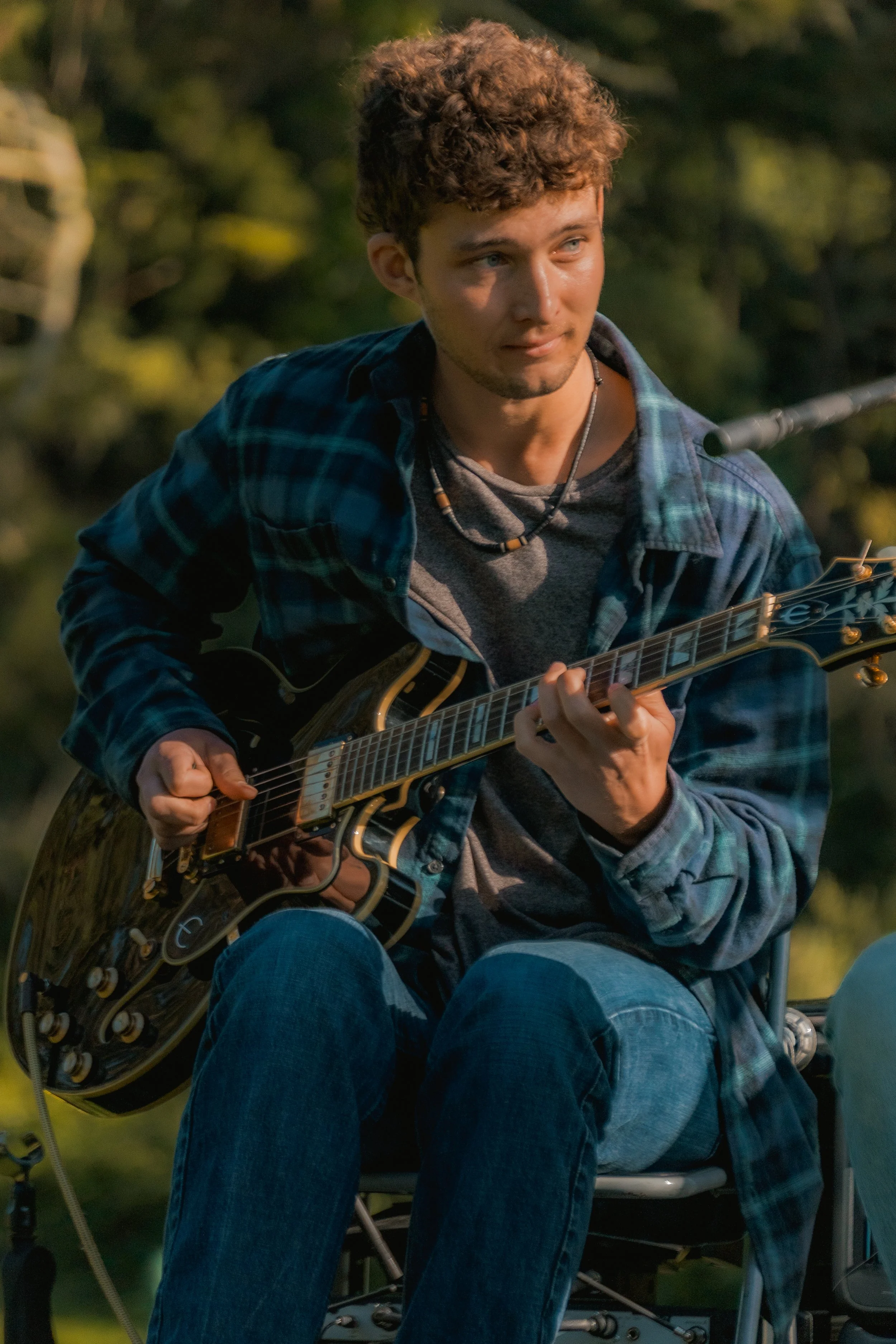 A young man with curly hair playing an electric guitar outdoors.