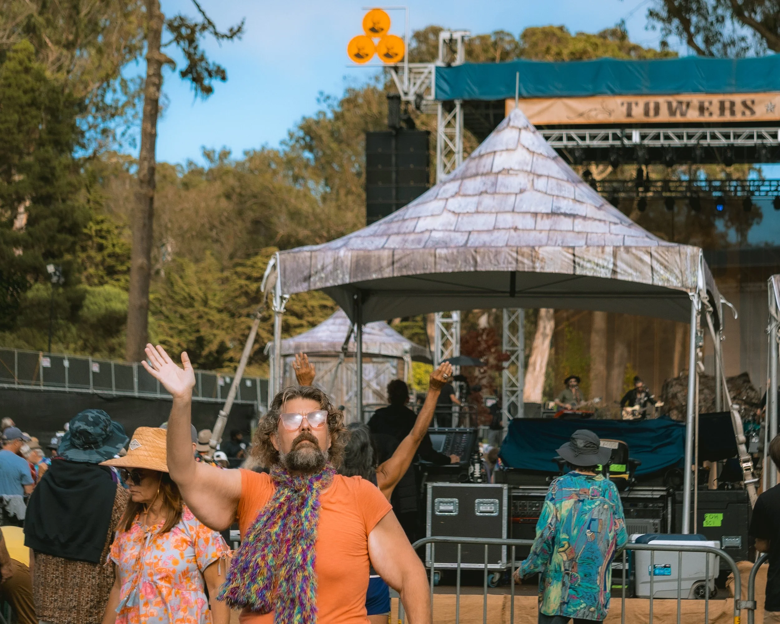 Man with long hair, beard, sunglasses, orange shirt, and colorful scarf raising arms in front of an outdoor stage with a band and audience