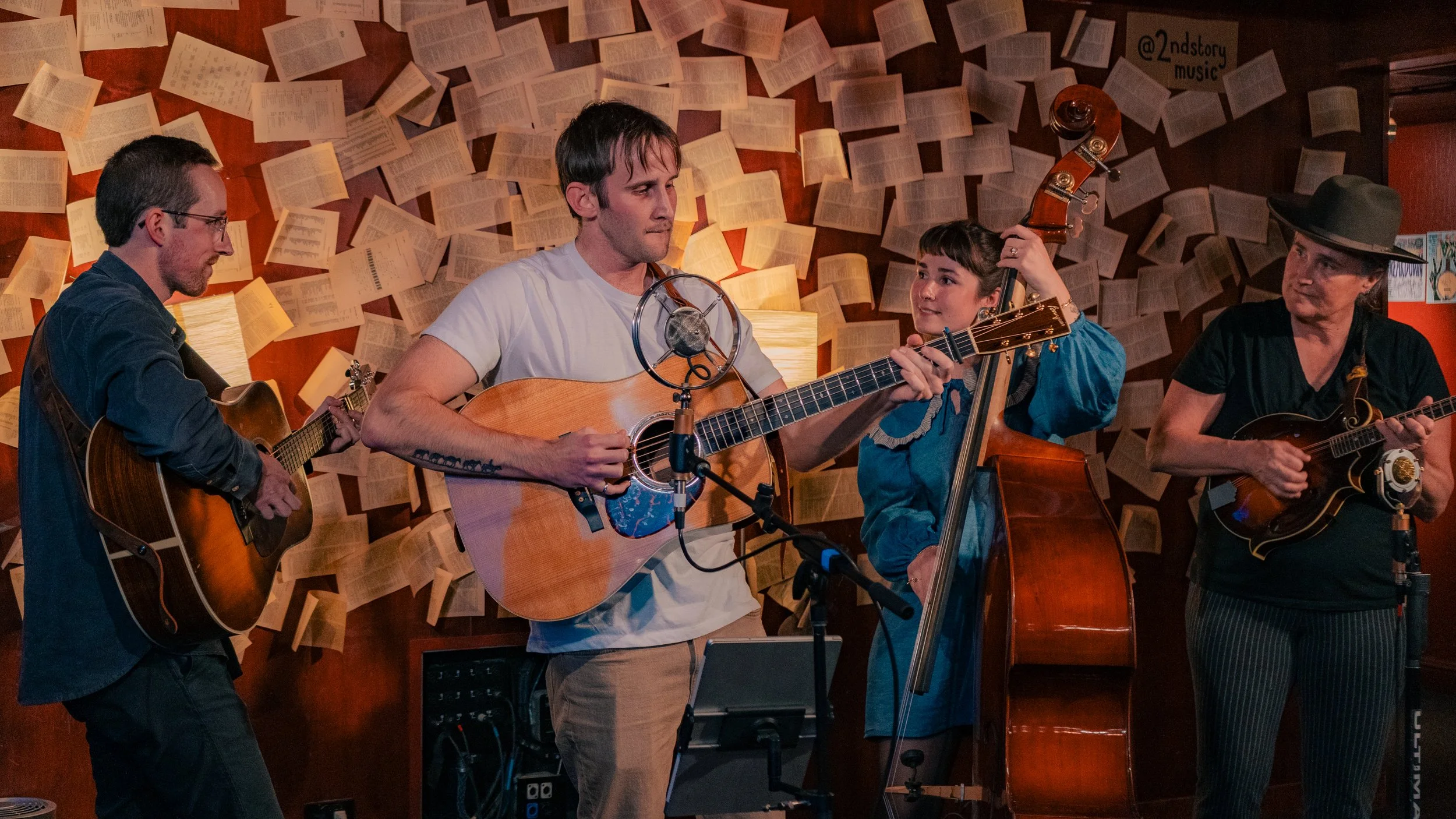 Four musicians performing with guitars and a double bass in a cozy, wood-paneled room decorated with pages of sheet music on the wall.