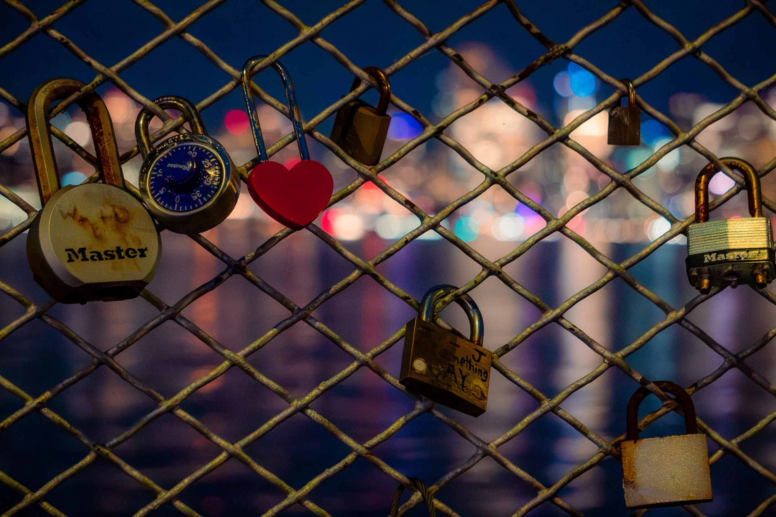 Locks attached to a metal fence at night with city lights in the background, including a round lock labeled 'Master', a red heart-shaped lock, and other locks, one with graffiti.