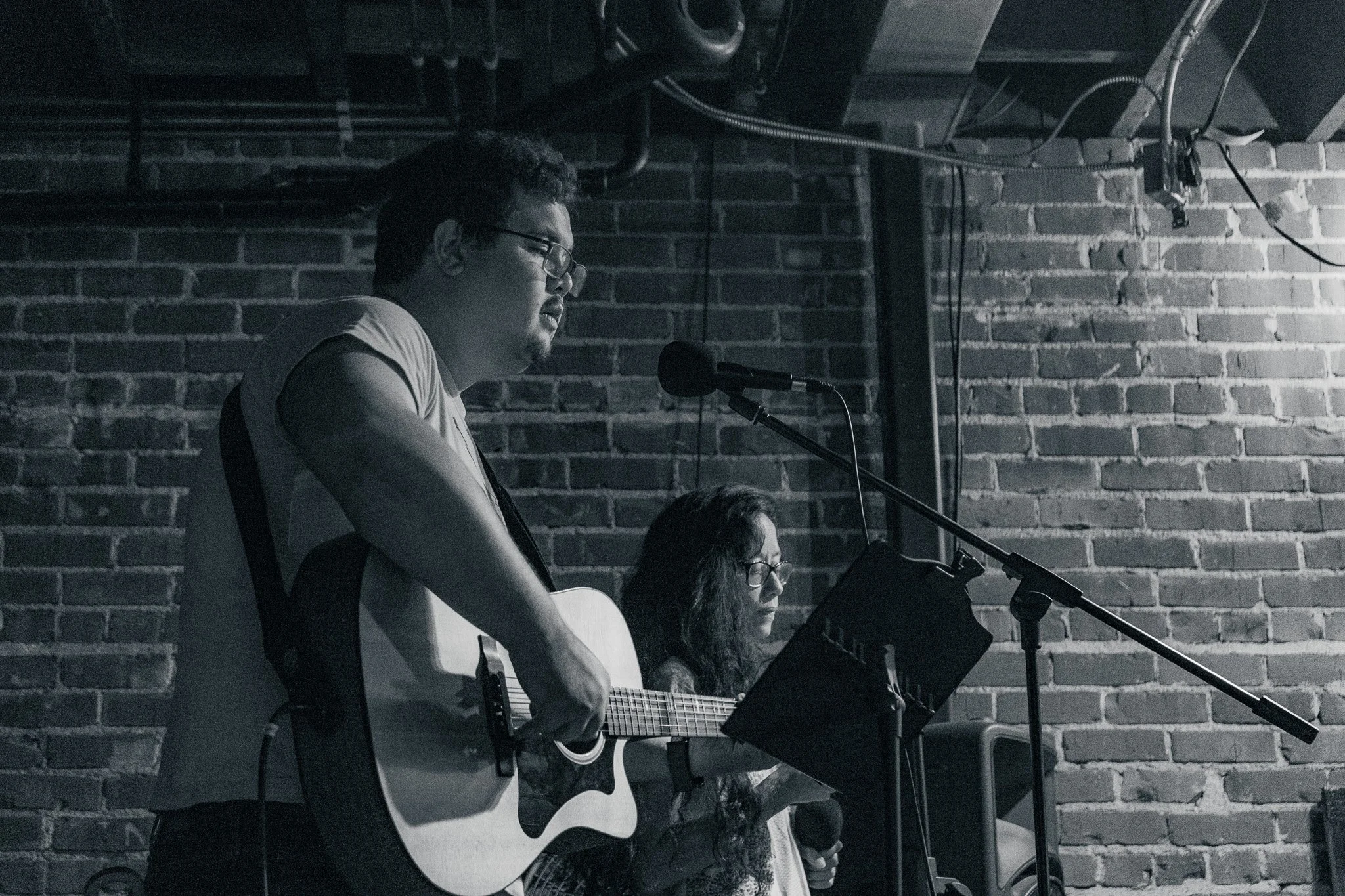 A man and a woman performing music on stage in front of a brick wall, with the man playing an acoustic guitar and the woman holding a microphone.