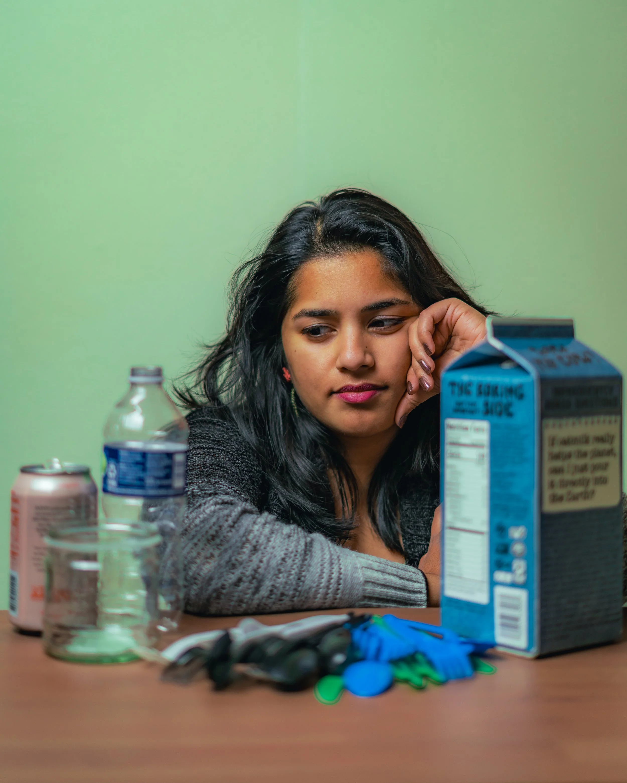 A young woman with long dark hair resting her head on her hand at a table with empty bottles, cans, a glass, a box of cereal, and scattered poker chips.