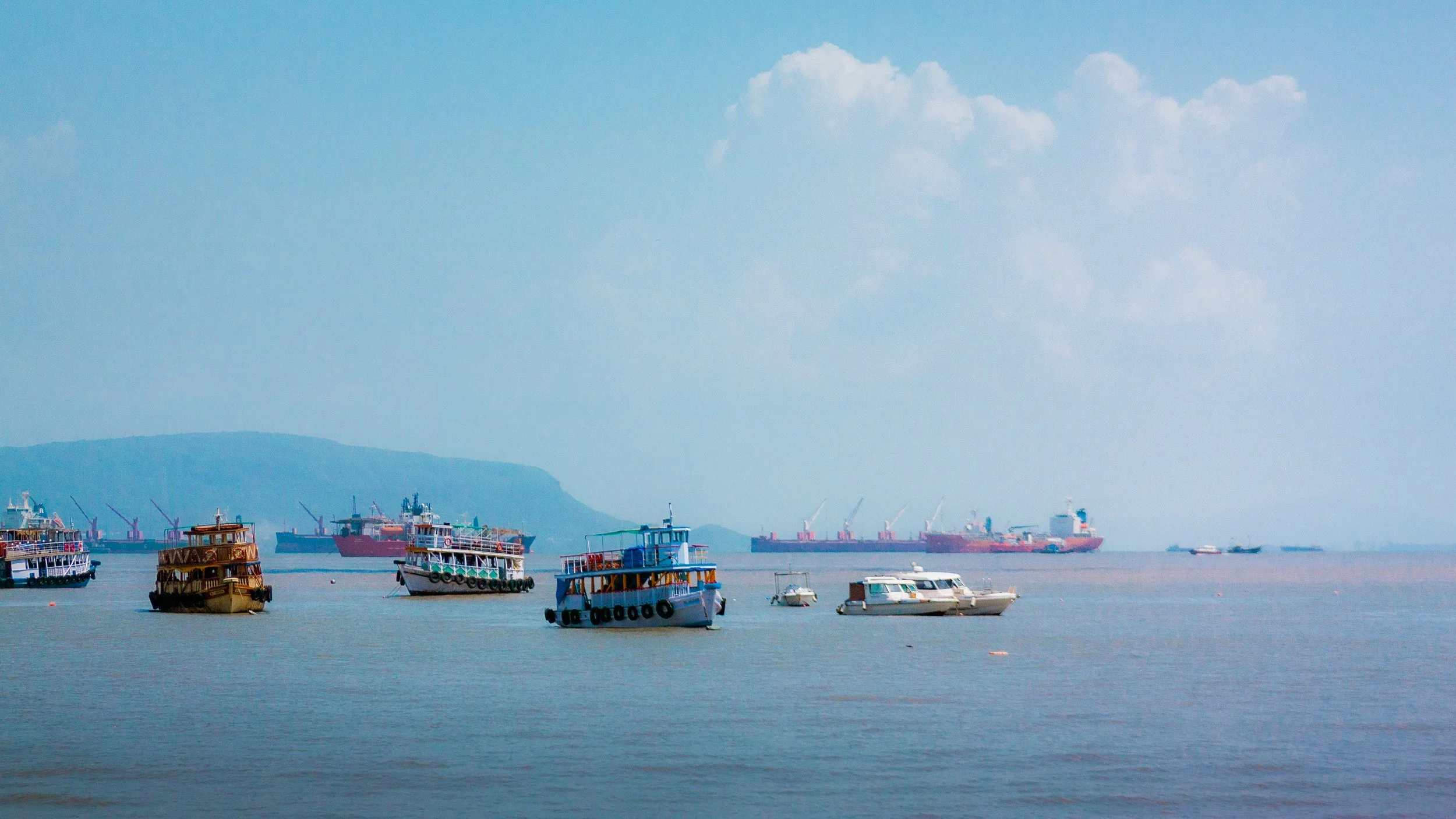 Multiple boats and ships on calm water with a distant shoreline and partly cloudy sky.