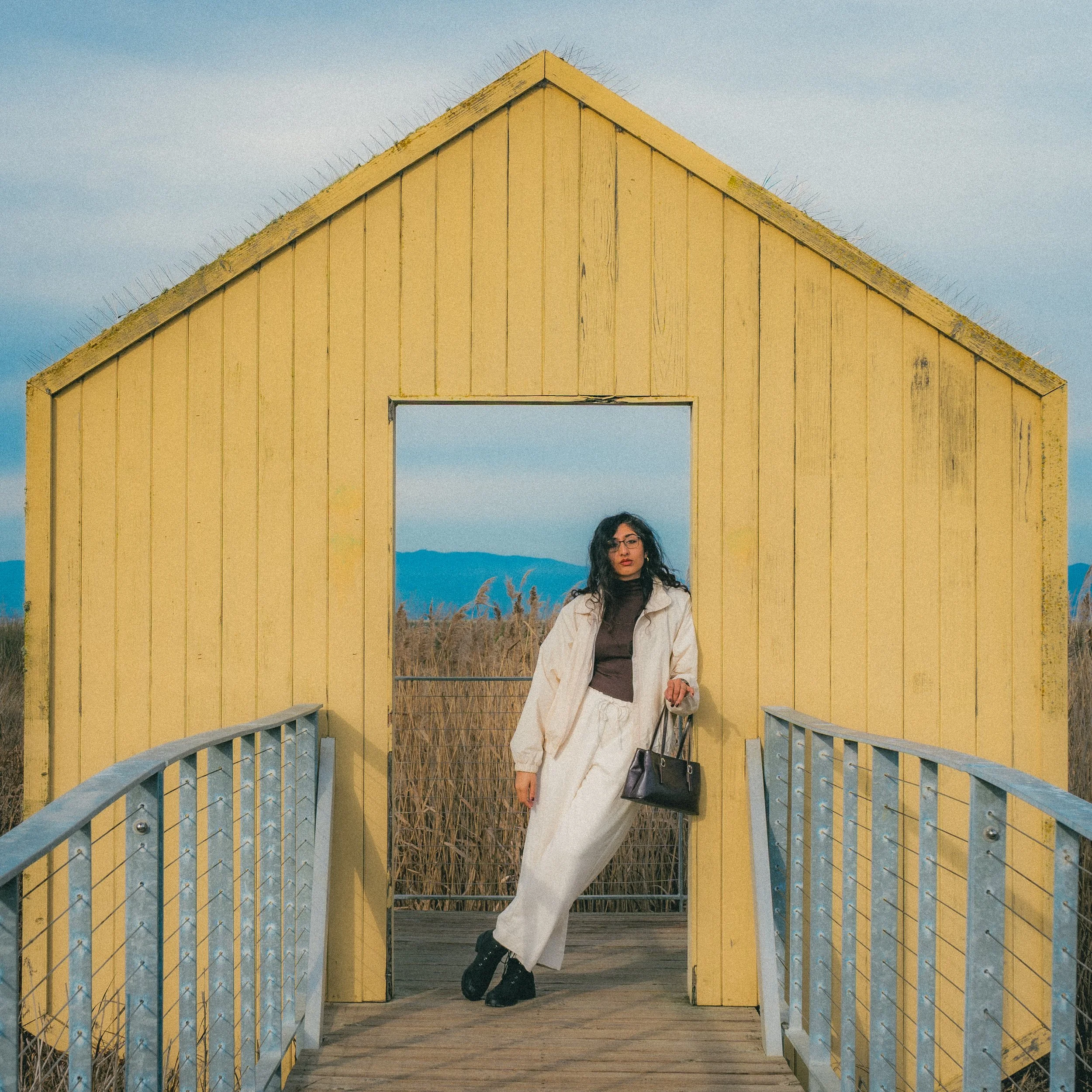 A woman standing in a doorway of a yellow wooden structure on a bridge, with a landscape of tall grass and mountains in the background.