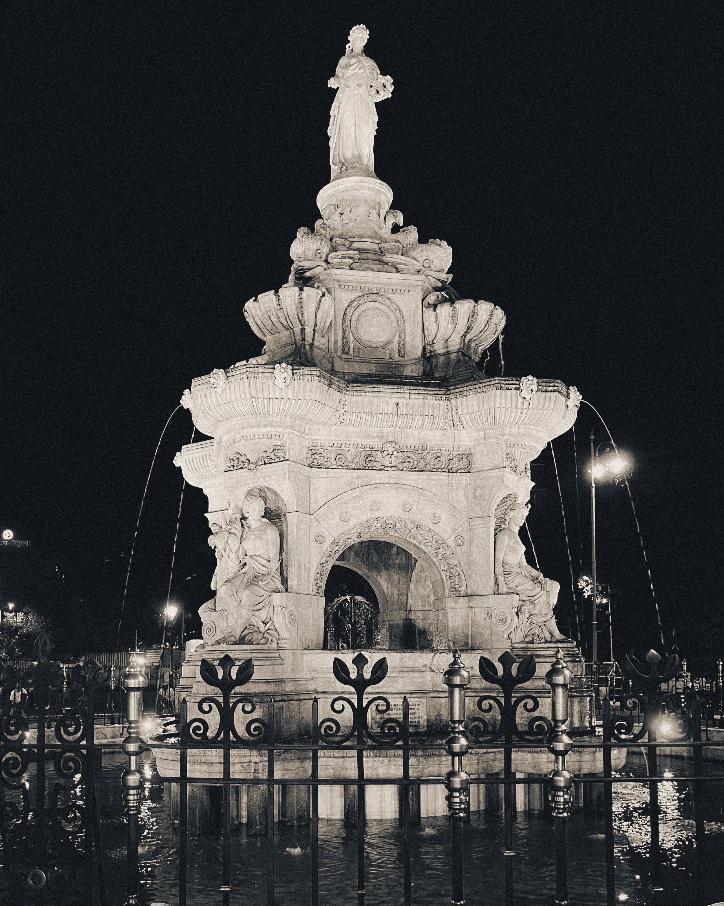 Nighttime view of an ornate, historic fountain with sculptures and water spouts, illuminated against a dark sky.
