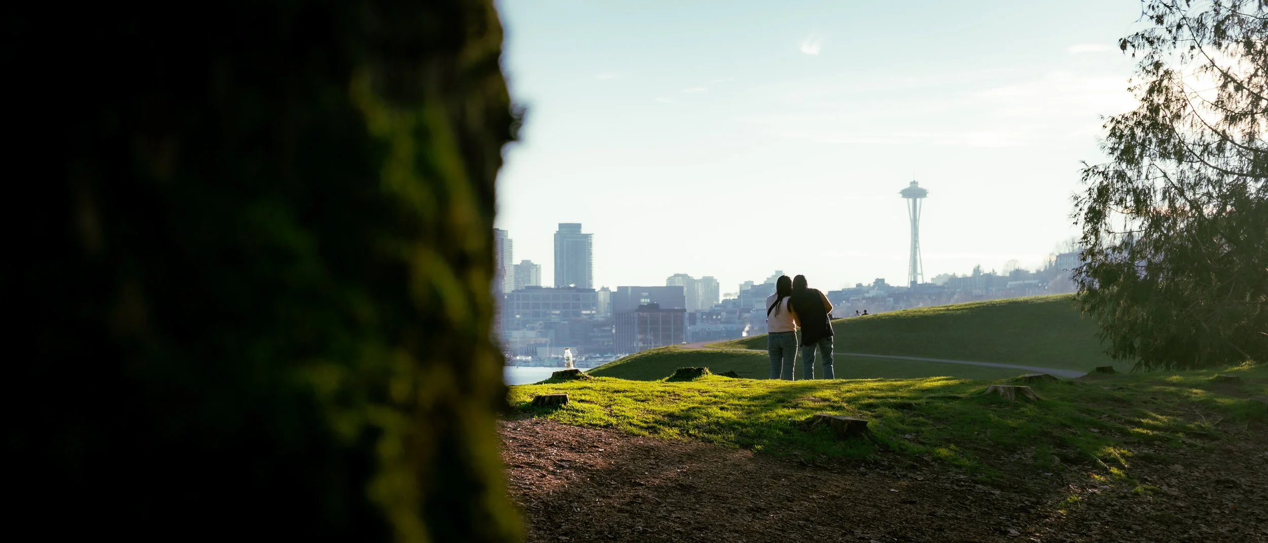 Two people walking together in a park with a city skyline, including the Space Needle, in the background on a sunny day.