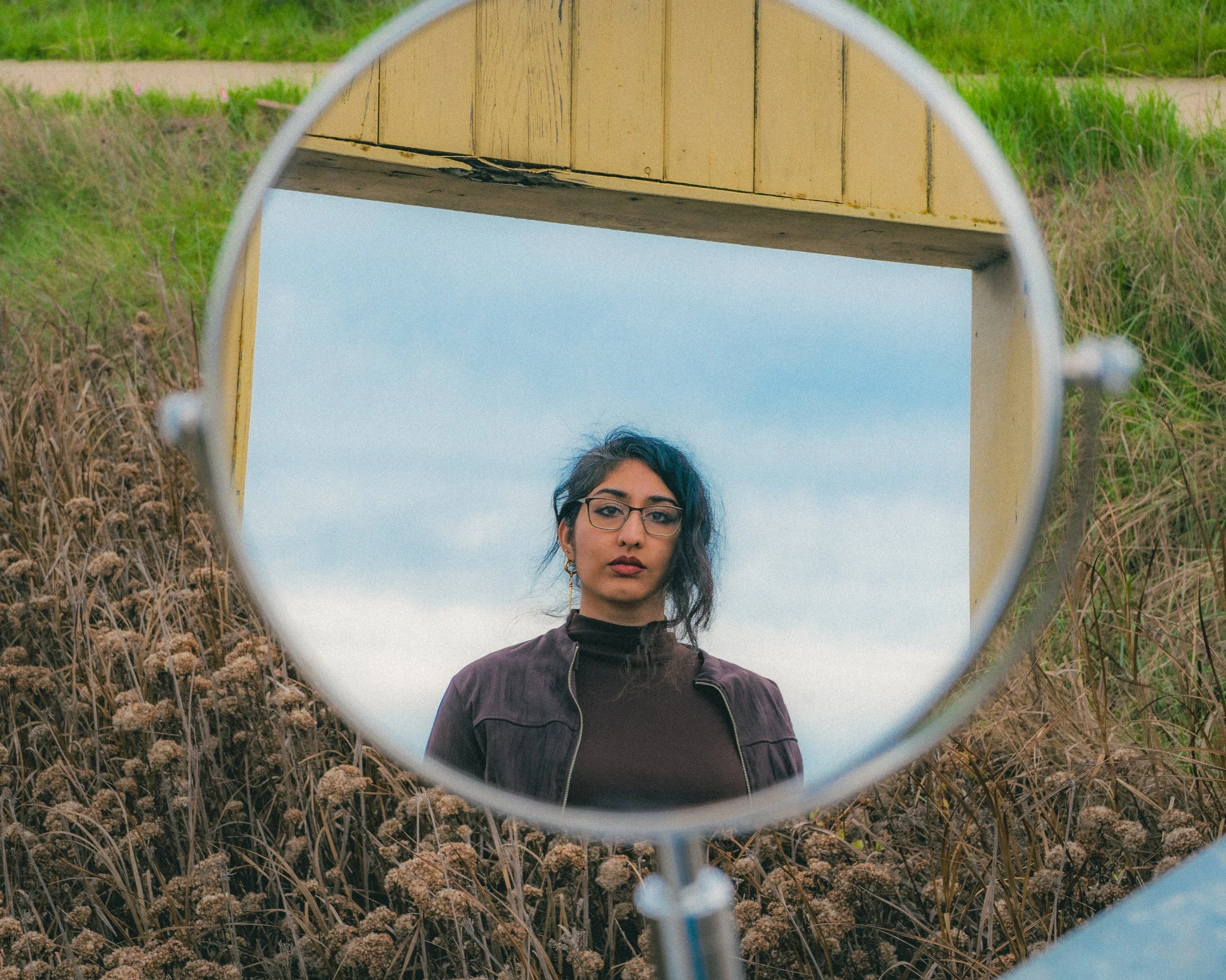 A woman with glasses and dark hair looking into a mirror outdoors, with tall dried grass and green hills in the background.
