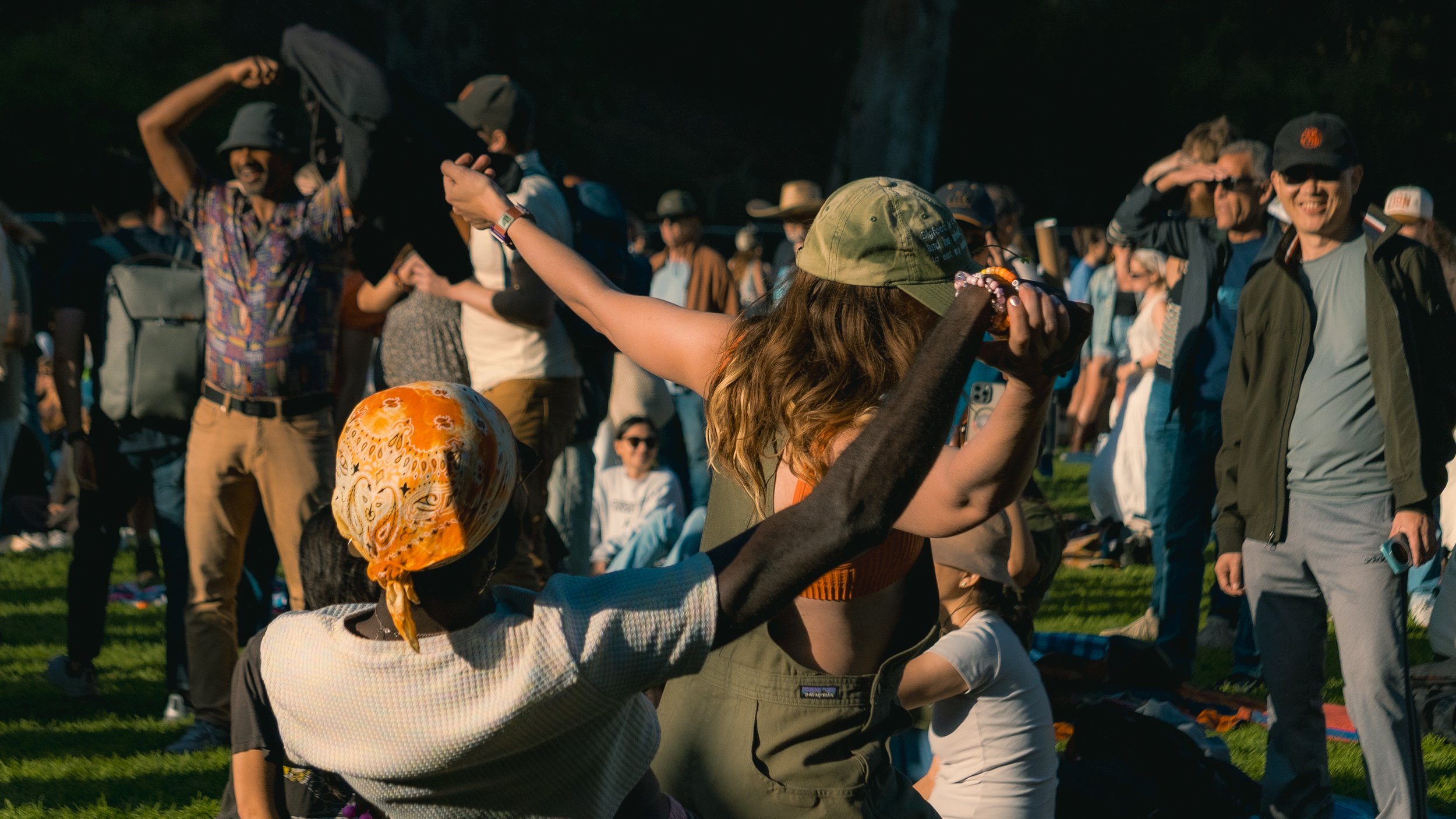 People gathered outdoors in a park or field during the day, some standing, some sitting, with a woman in the foreground wearing a green cap and a woman wearing a colorful headscarf dancing or celebrating in front, amidst a diverse crowd.