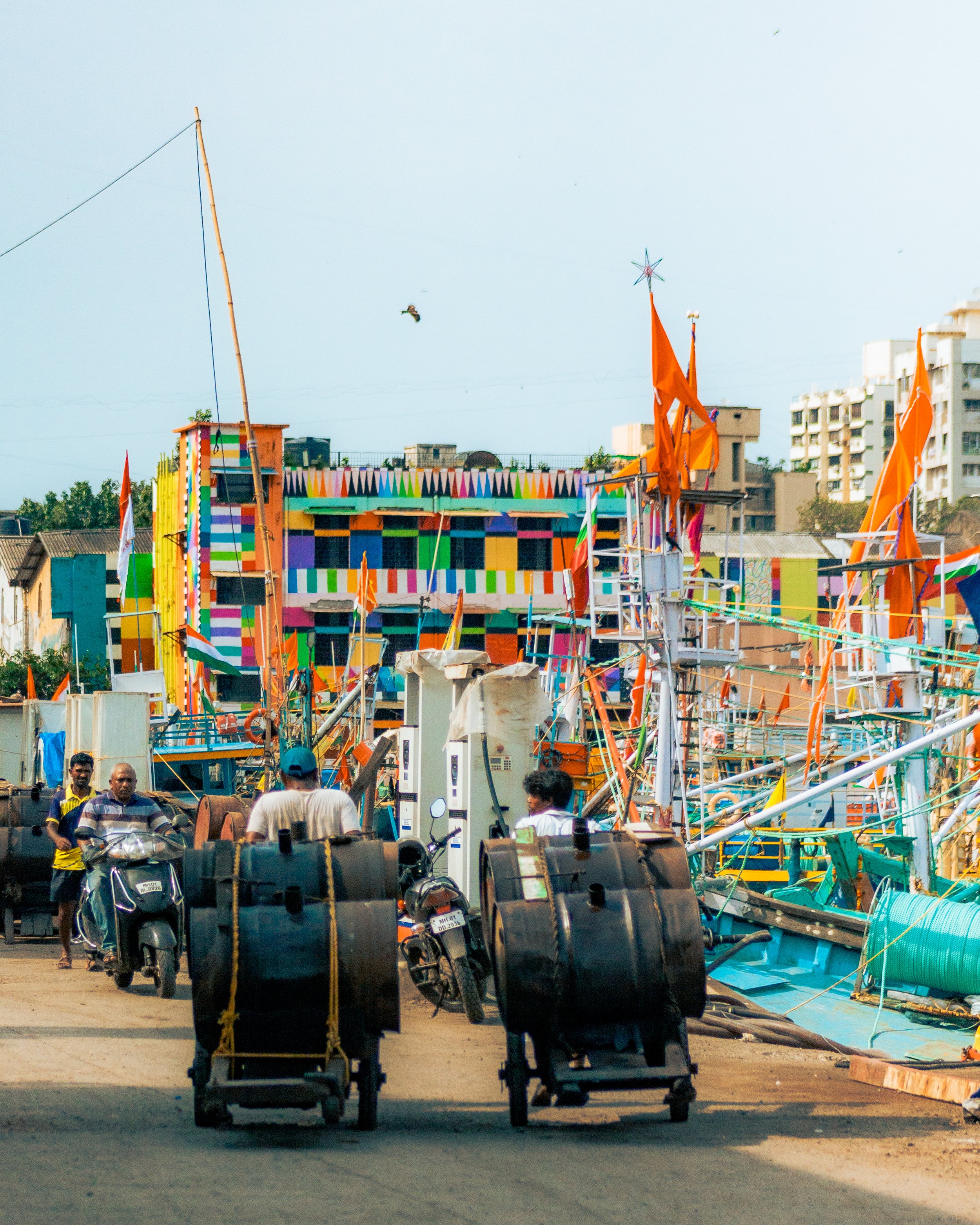 People walking near colorful boats and flags on a dock in a coastal city with high-rise buildings in the background.