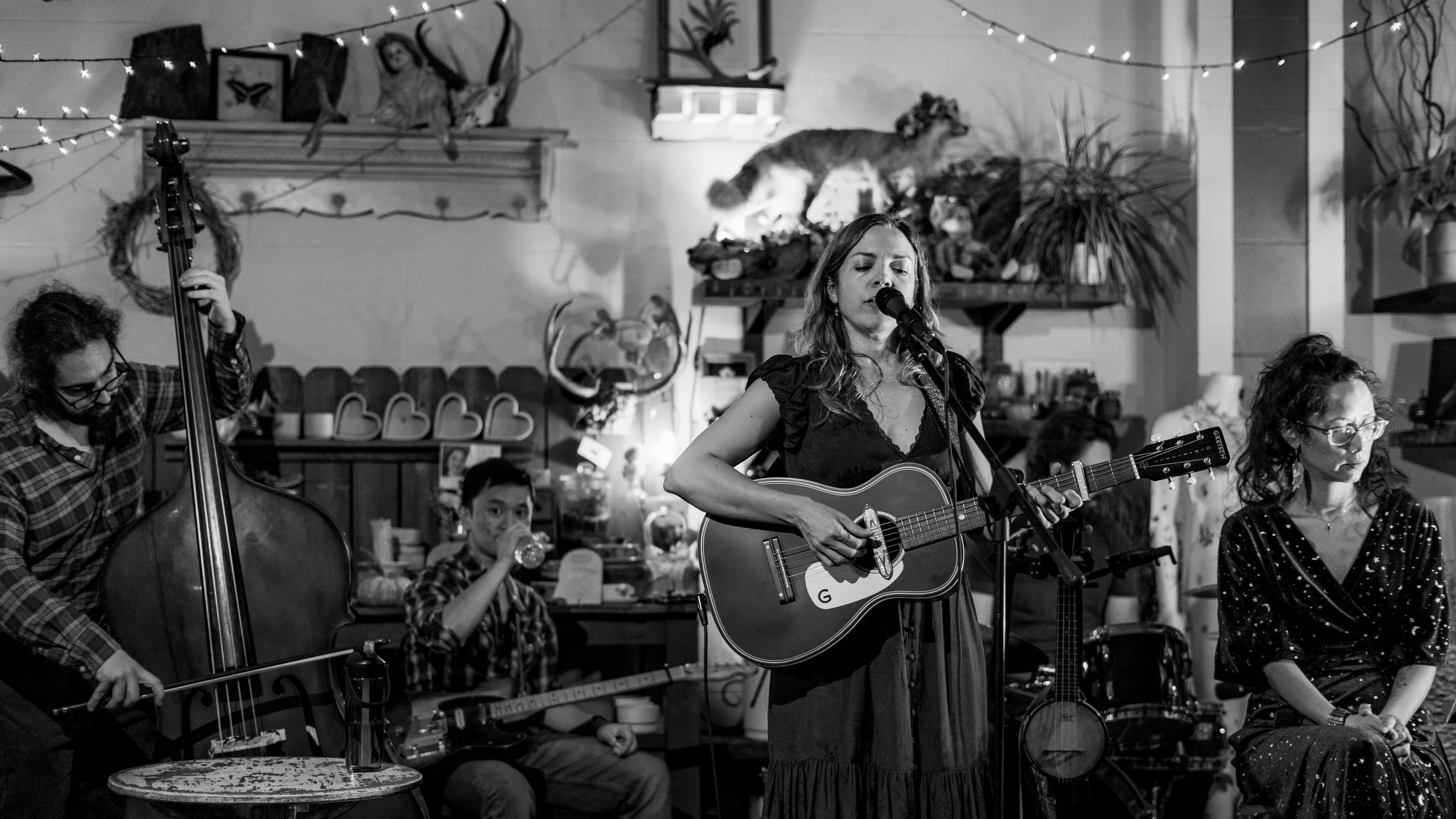 A black and white photo of a musical performance in a cozy, decorated room. A woman is singing and playing an acoustic guitar into a microphone. To her left, a person is playing a double bass, and behind the singer, a young boy is drinking from a gla