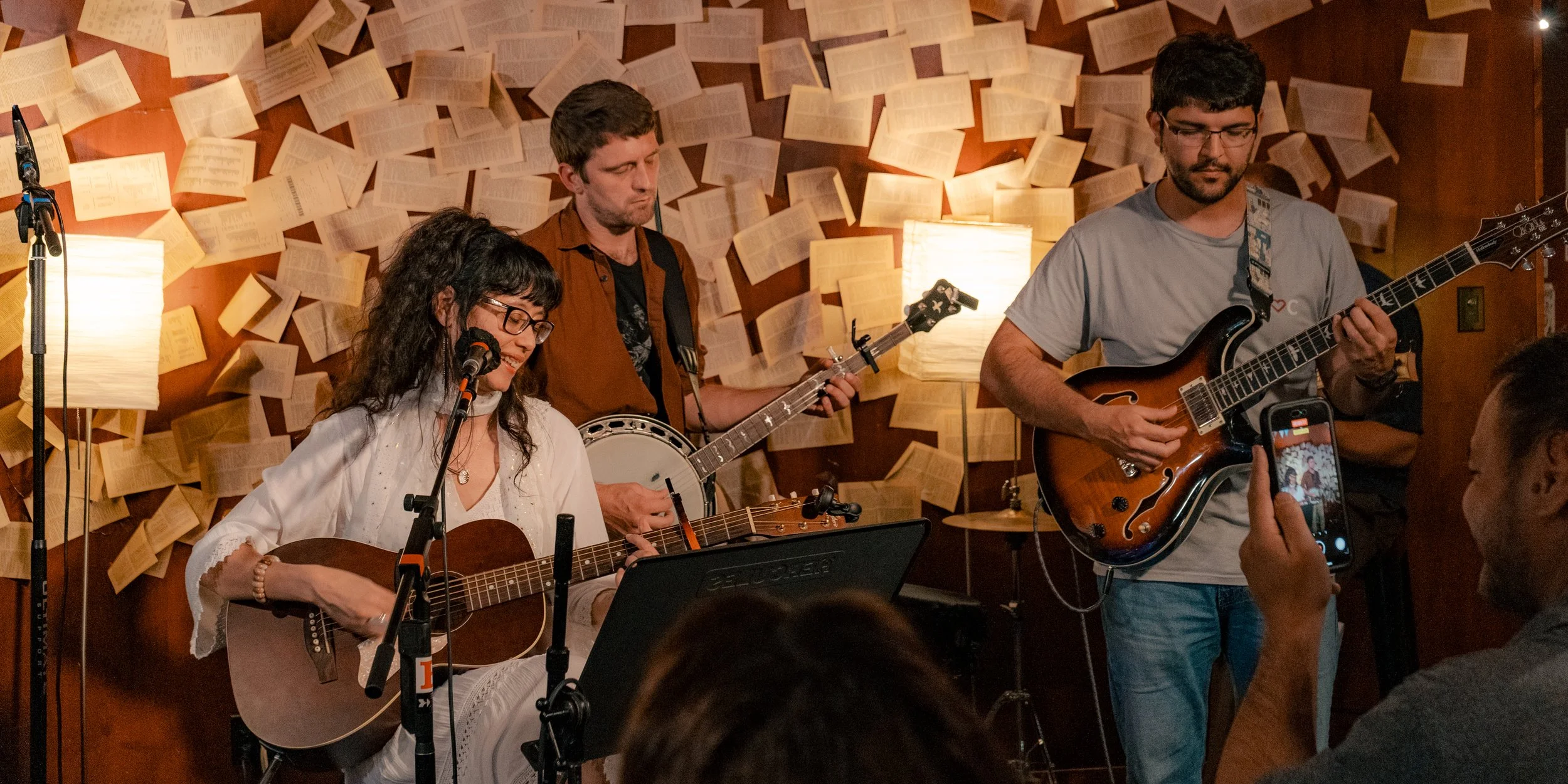 A musical group performing live in a cozy venue with a background of open books on the wall and warm lighting.