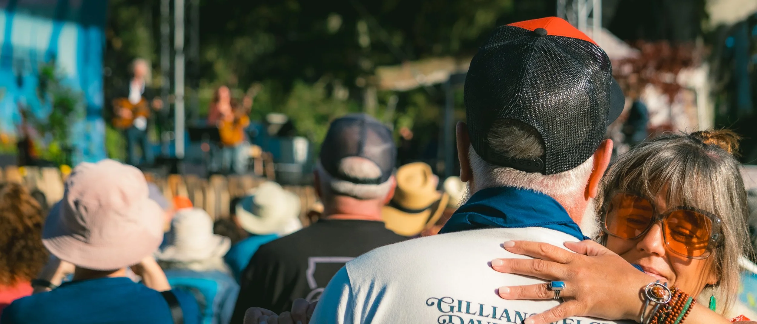 A crowd of people sitting outdoors watching a performance on a stage. The focus is on a woman with gray hair and large orange sunglasses hugging a man wearing a baseball cap with a red and black mesh back.