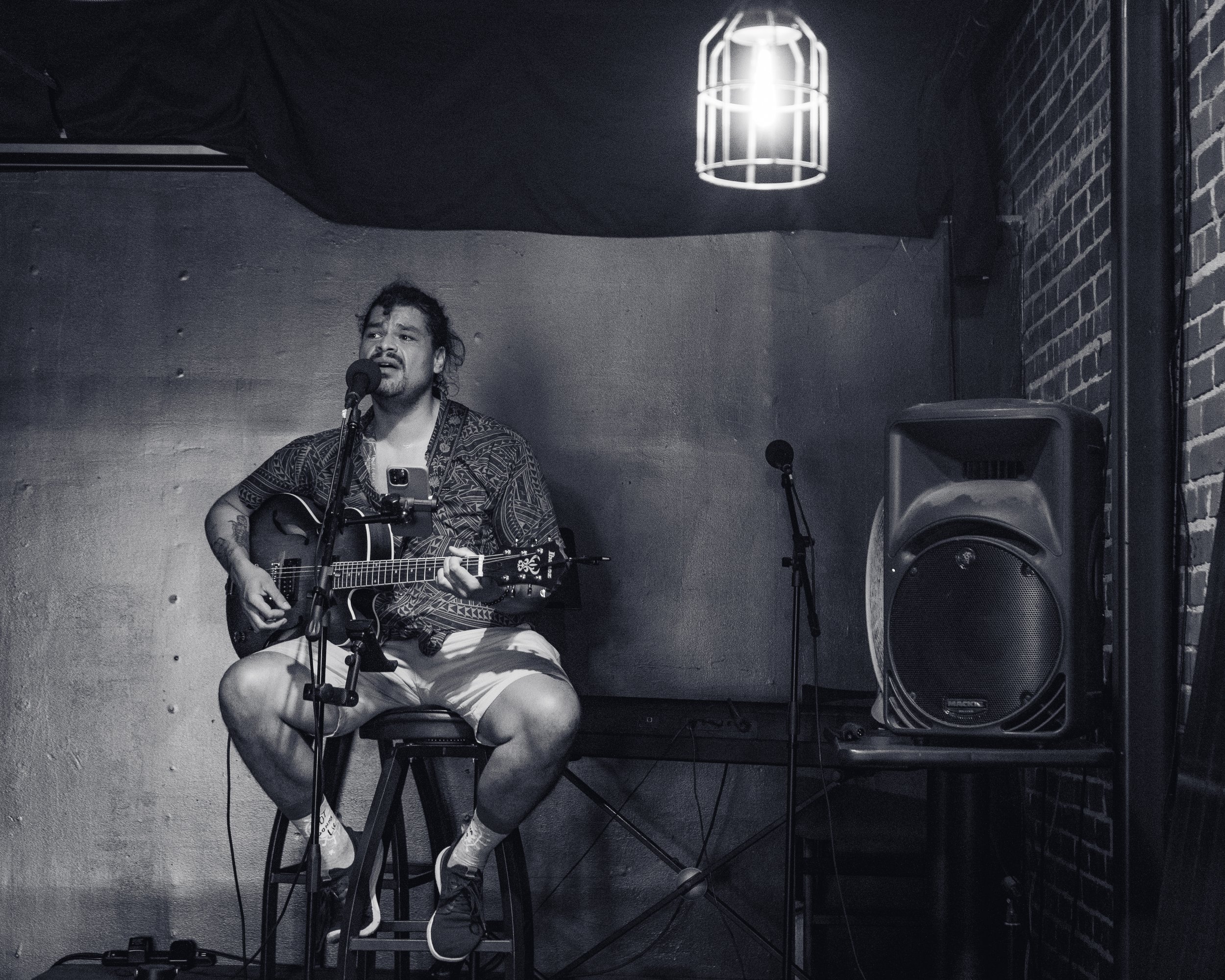 A man with a beard and curly hair, wearing a patterned shirt and shorts, is sitting on a stool playing an acoustic guitar and singing into a microphone in a small performance venue with a brick wall and a large speaker.