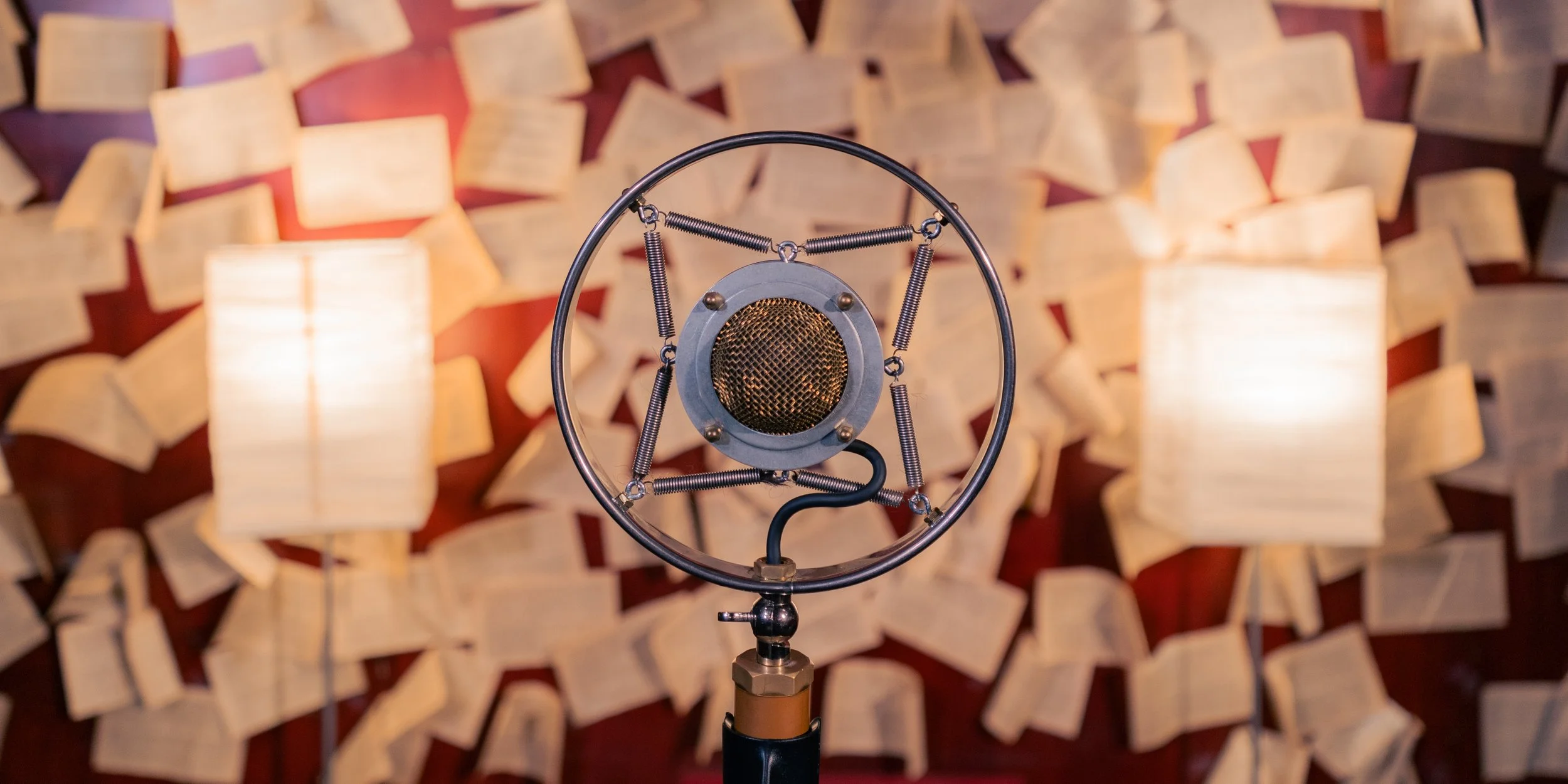 Vintage recording microphone held above open books scattered on a red surface, with two lamps providing warm lighting.