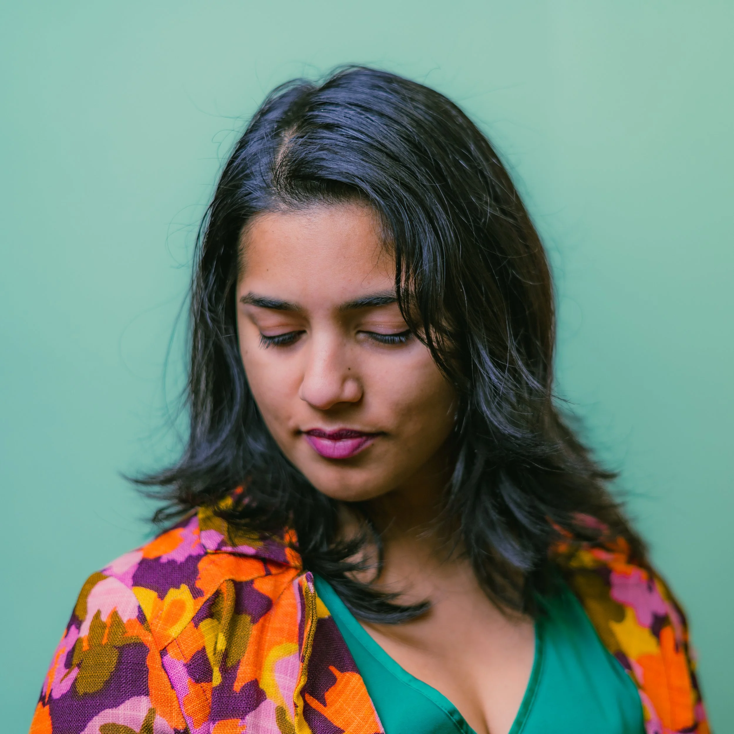 A woman with dark hair, closed eyes, wearing a colorful floral cardigan over a green top, against a turquoise background.