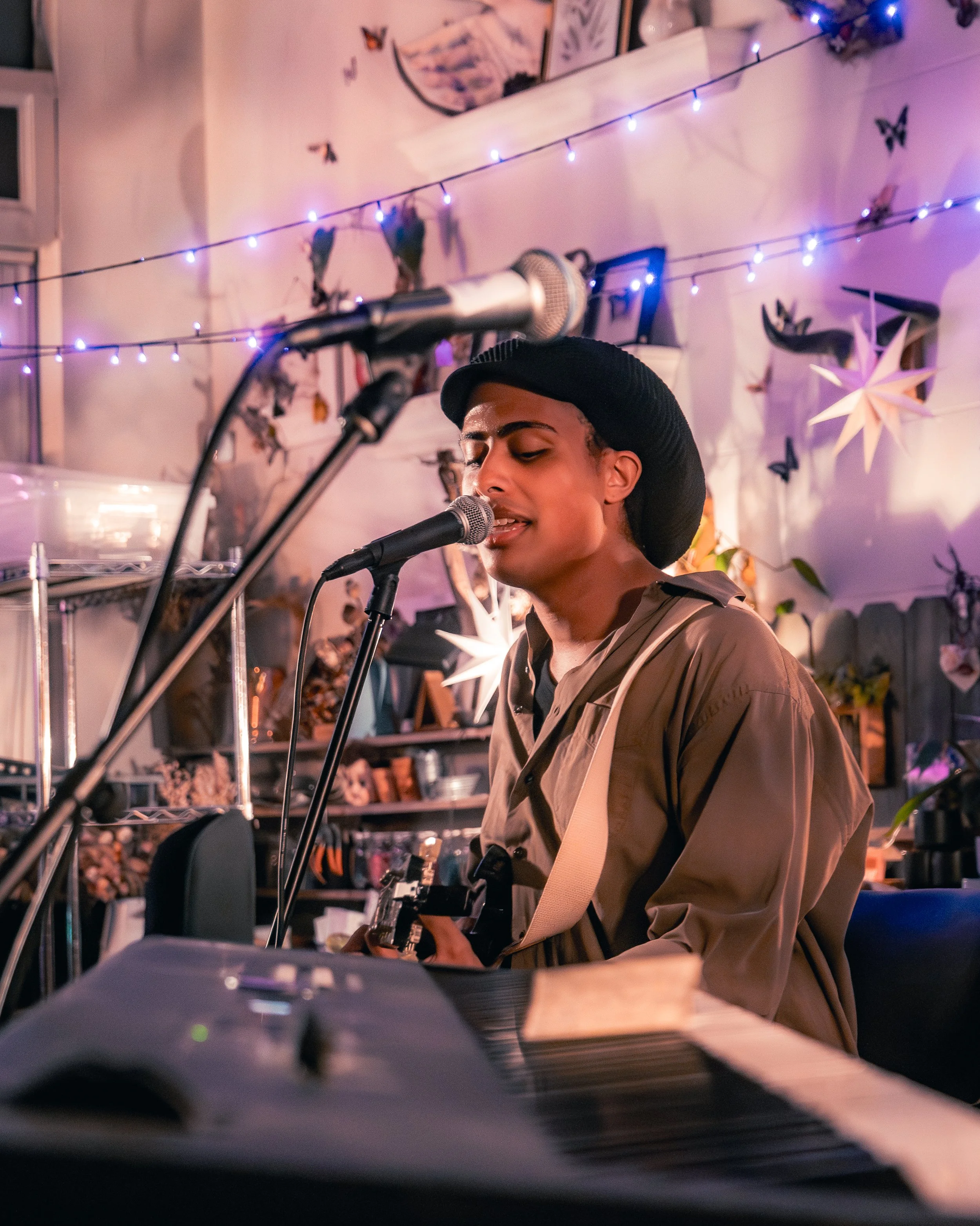 A young man with a hat singing into a microphone, playing guitar, surrounded by colorful fairy lights and decorative paper stars and butterflies in a cozy room.