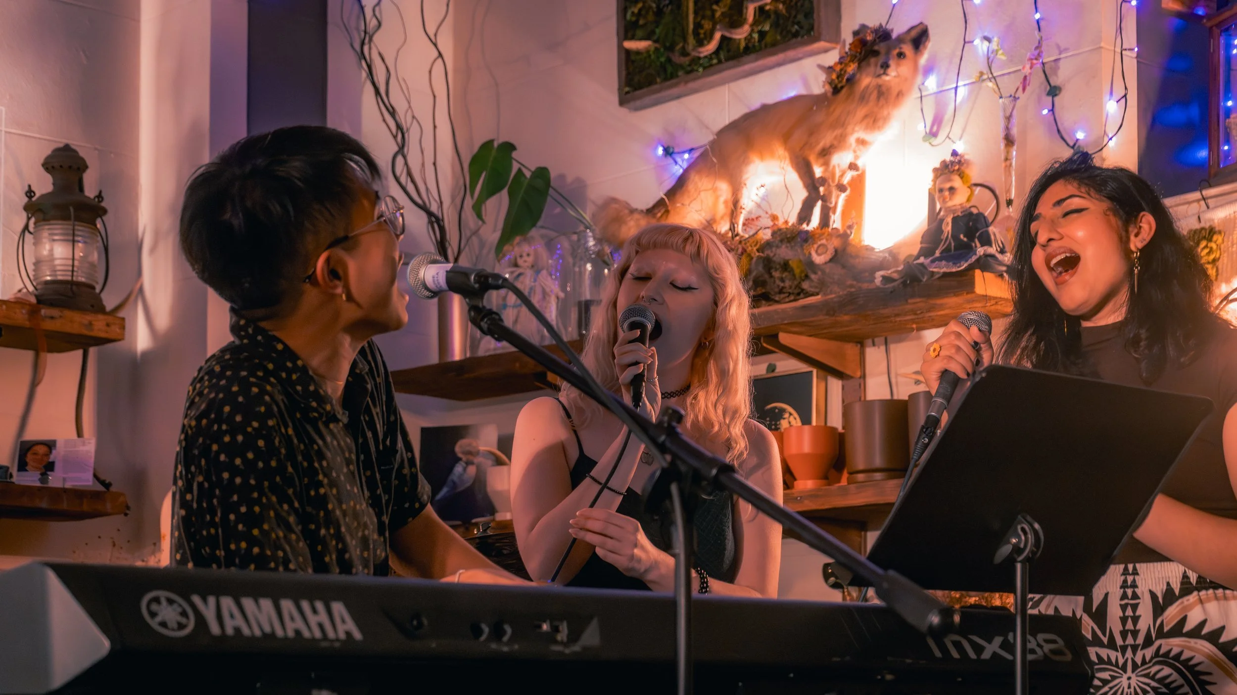 Three women singing and playing keyboard in a decorated room with fairy lights, shelves, and figurines.