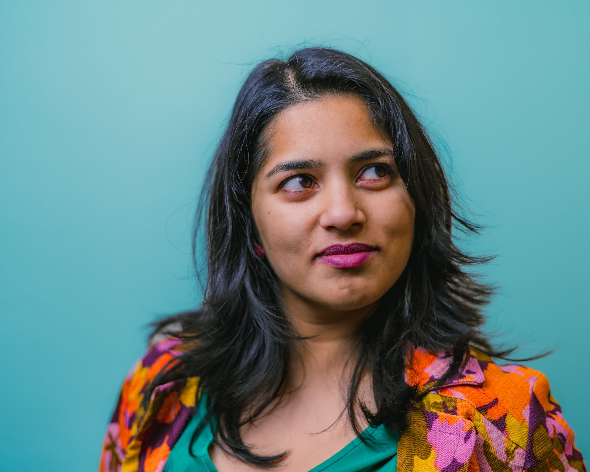 A woman with shoulder-length wavy black hair wearing a multicolored floral jacket and a green top, looking to her right against a teal background.