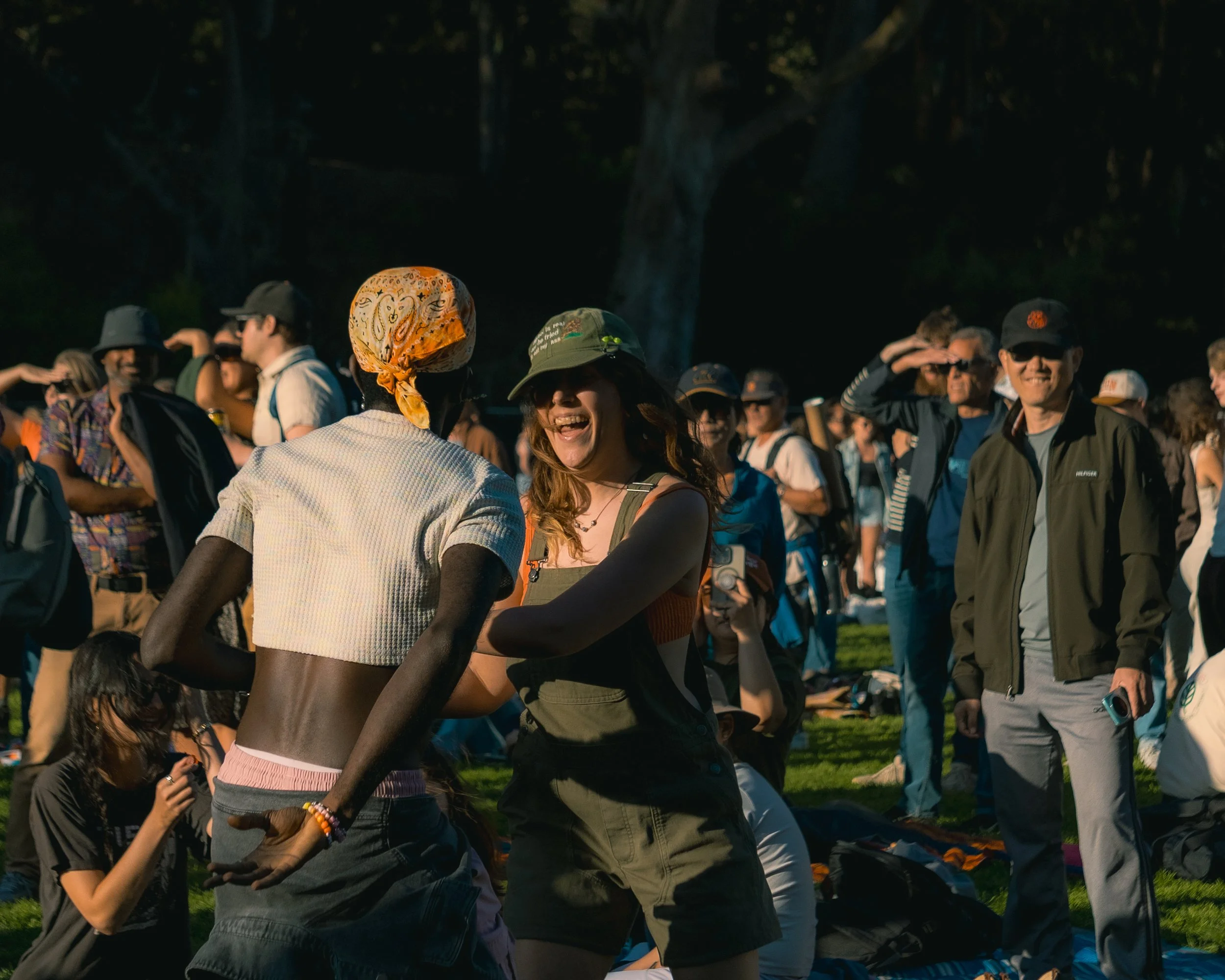 People outdoors, some dancing, some standing and smiling, in a park during daytime, with trees in the background.