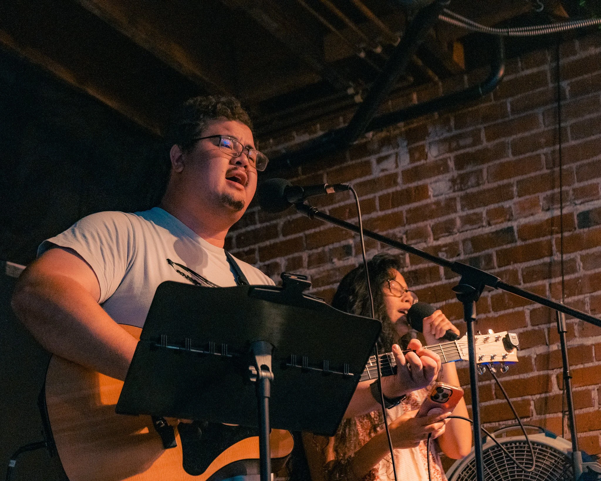 A man playing an acoustic guitar and singing into a microphone on stage with a woman singing into a microphone and looking at her phone, against a brick wall background.