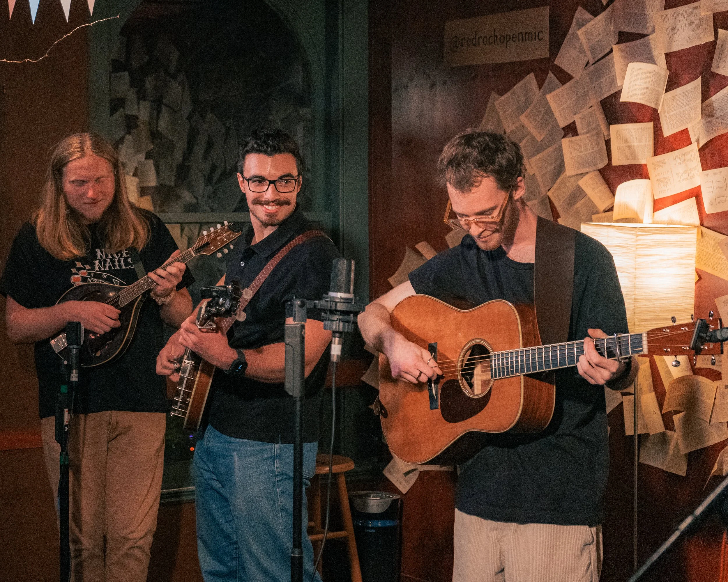 Three musicians playing instruments in a cozy room with pages of books on the wall and a lamp, during a live music performance.