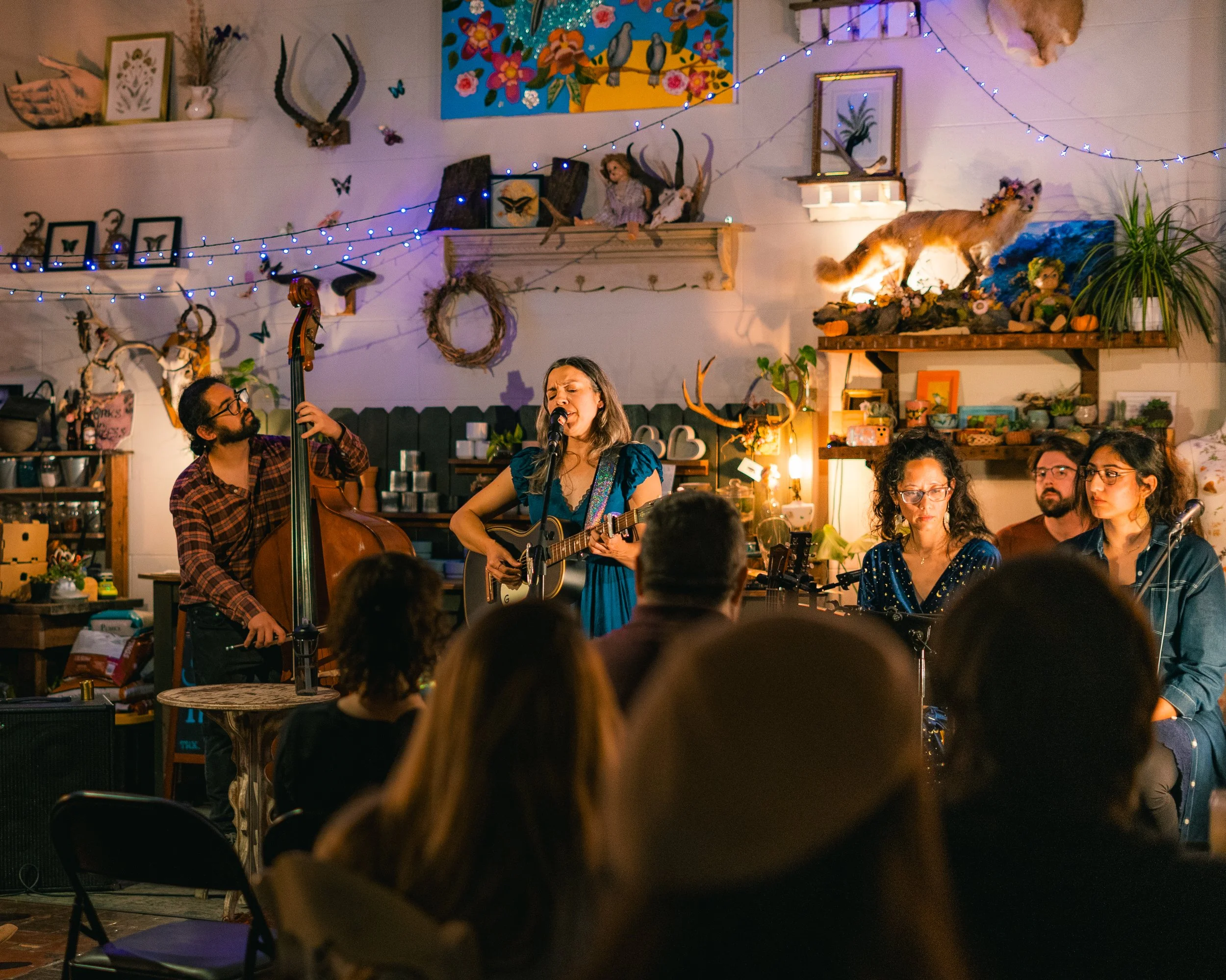 A band performs live music in a cozy, decorated indoor space with an audience watching. The backdrop features shelves with plants, framed pictures, deer antlers, and festive decorations including string lights and mounted animals.