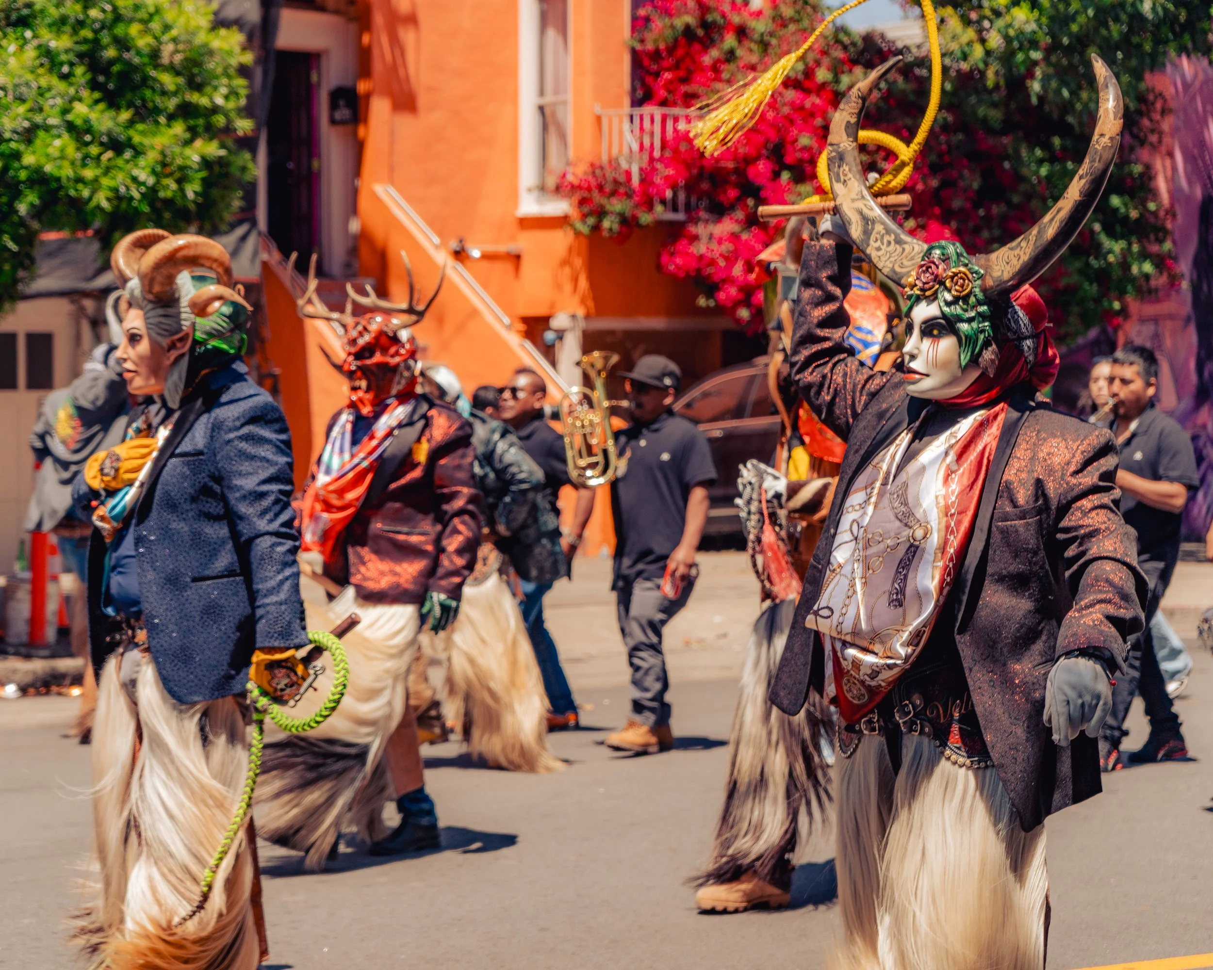 Participants in a cultural parade dressed in elaborate costumes with masks, horns, and fur, walking down a street with colorful buildings and spectators in the background.