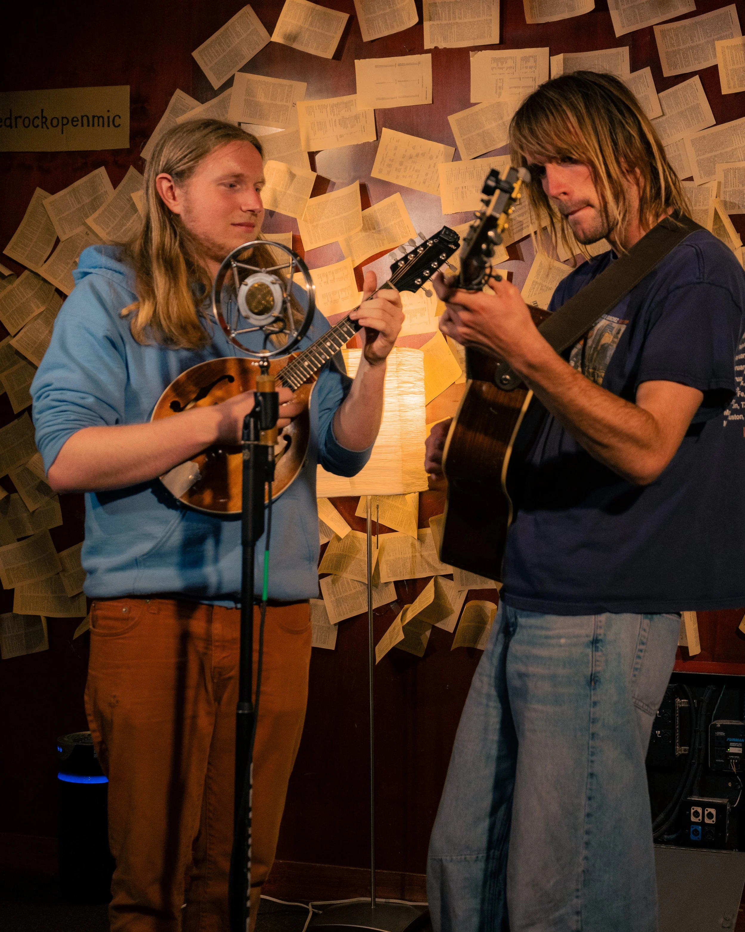 Two young men playing guitars and singing into a vintage microphone in a room decorated with open books on the wall and a lamp in the background.