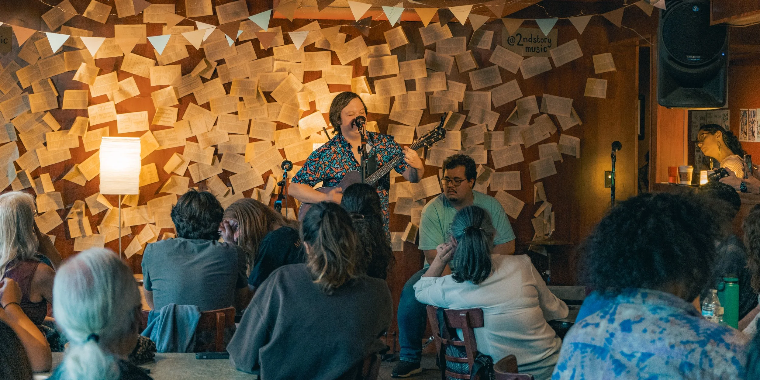 A live music performance at a cozy venue with a wooden wall decorated with open book pages and a string of banners. A male guitarist and singer is performing, surrounded by an audience seated at tables, some enjoying drinks. There are also stage ligh