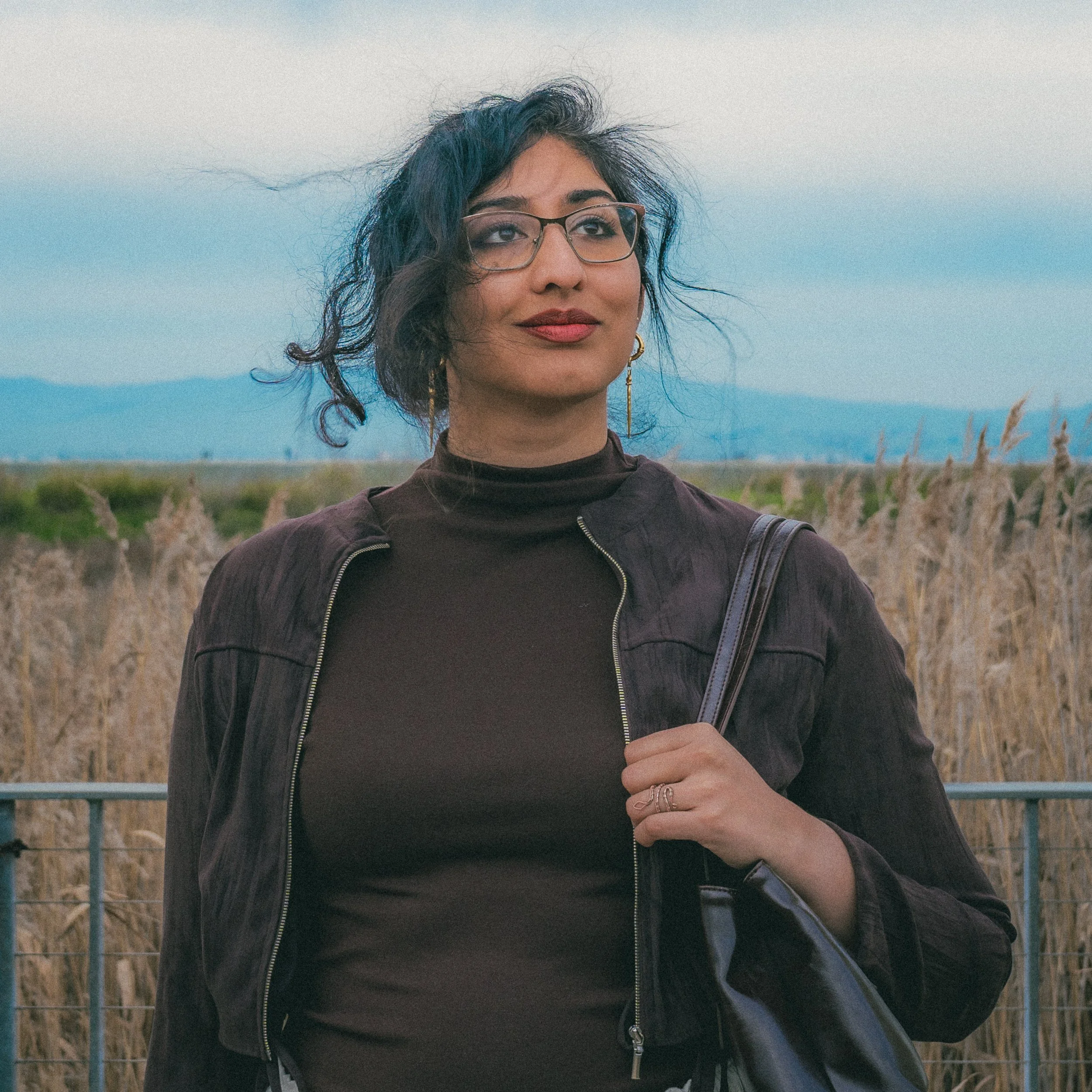 A woman with black, curly hair and glasses standing outdoors near a field with dry grass, mountains in the distance, and a cloudy sky.
