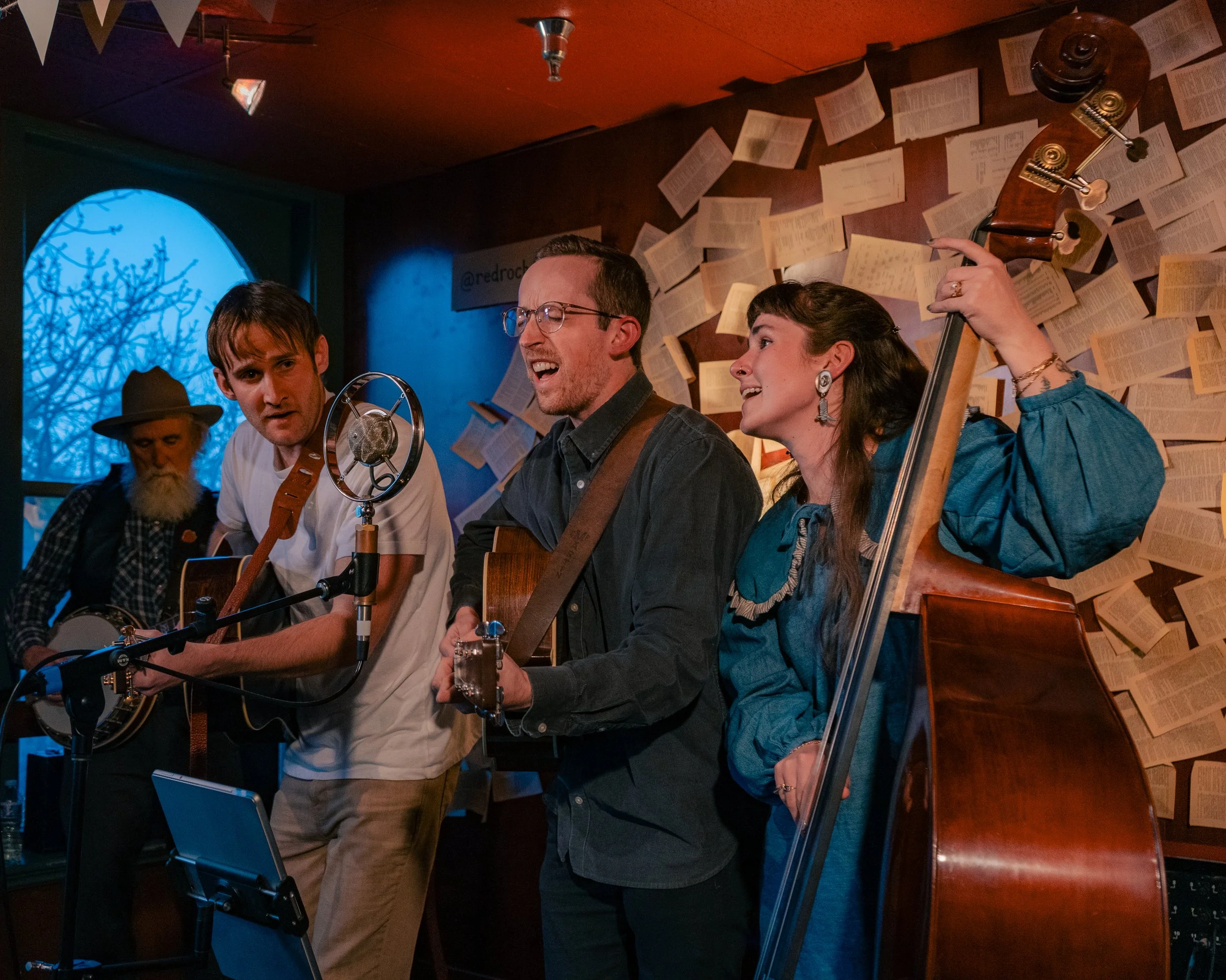 Four musicians perform together indoors, with newspaper pages on the wall and a window showing a blue sky with tree branches. They are playing guitars, a banjo, and a double bass, with a vintage microphone in front of them.