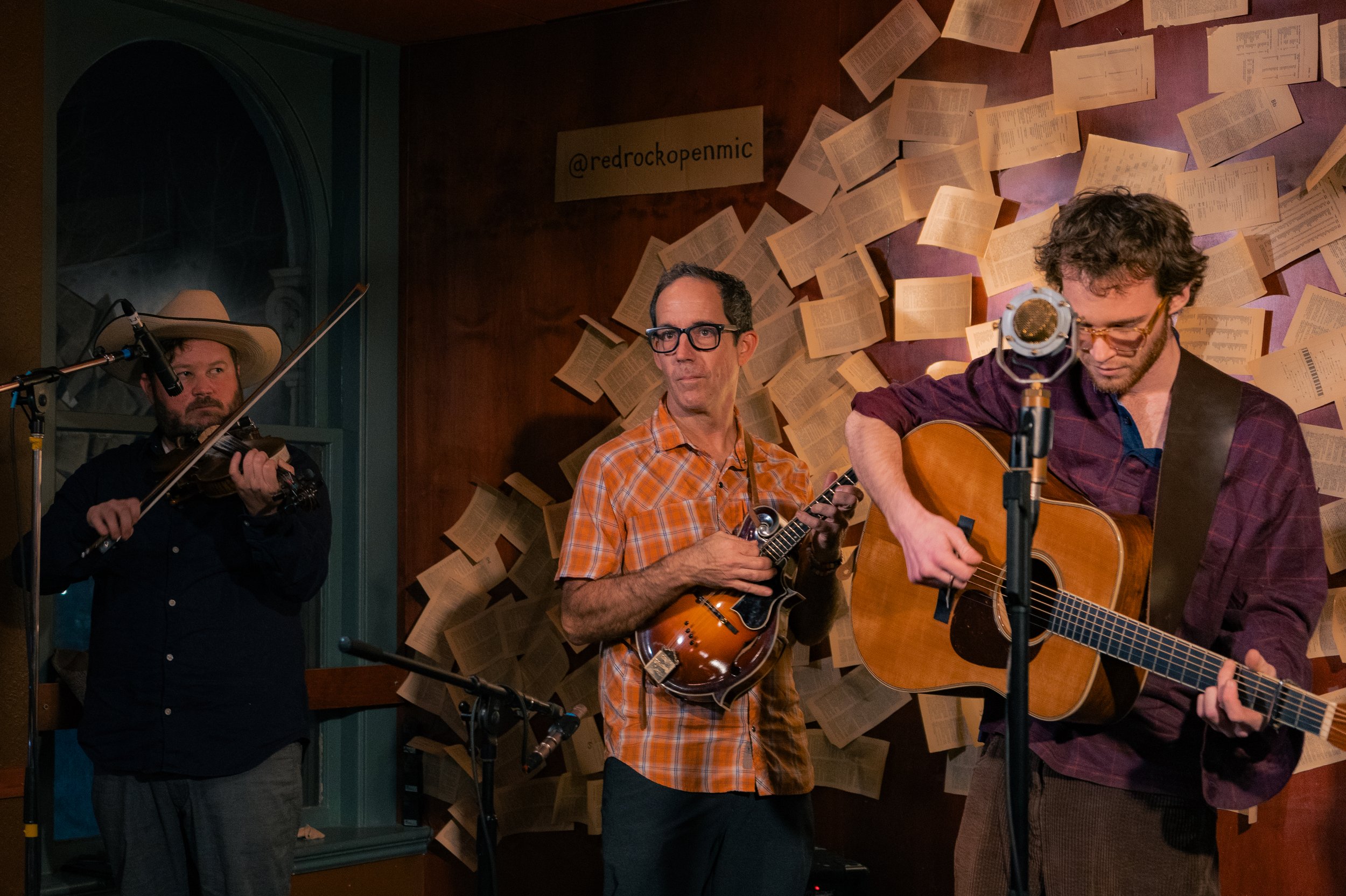 Three musicians playing instruments in a room with background wall covered in open books and a sign that reads '@redrockopenmic'.