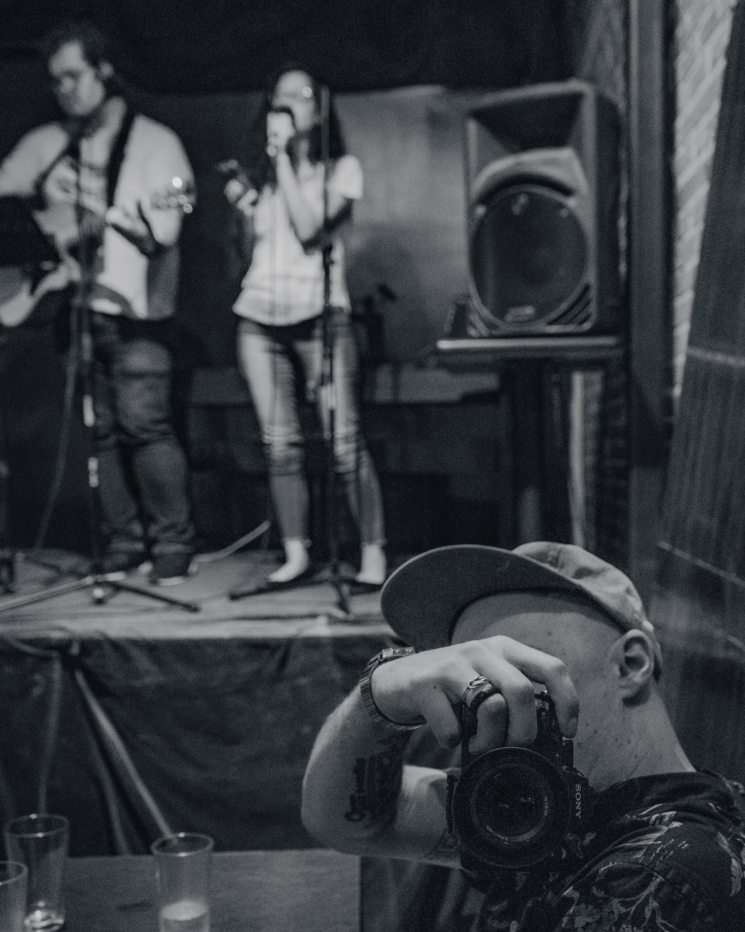 A person taking a photo of a live music performance in a dimly lit venue, with musicians playing guitar and singing on stage in the background.