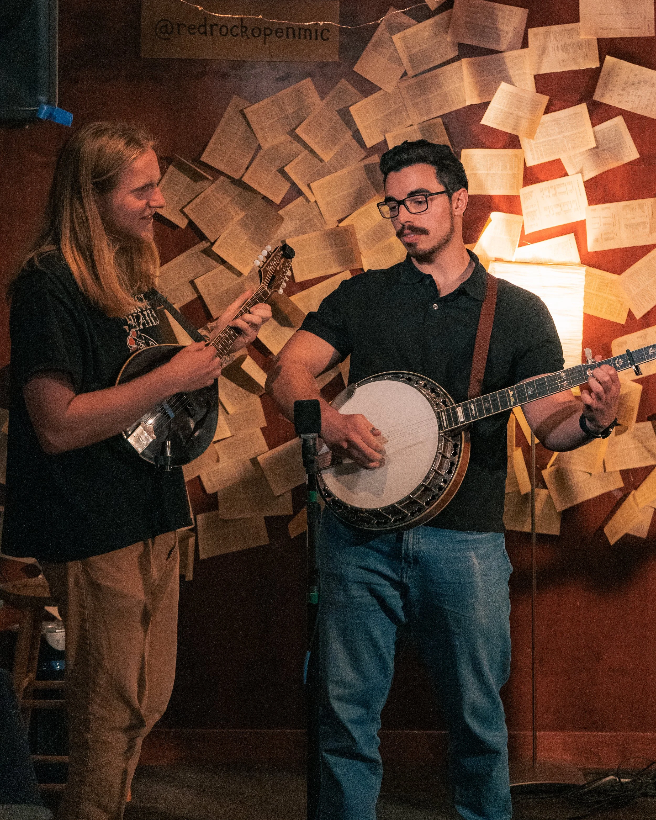 Two musicians, a woman and a man, playing guitars and singing into a microphone in a dimly lit bar or lounge with a wall decorated with open books.